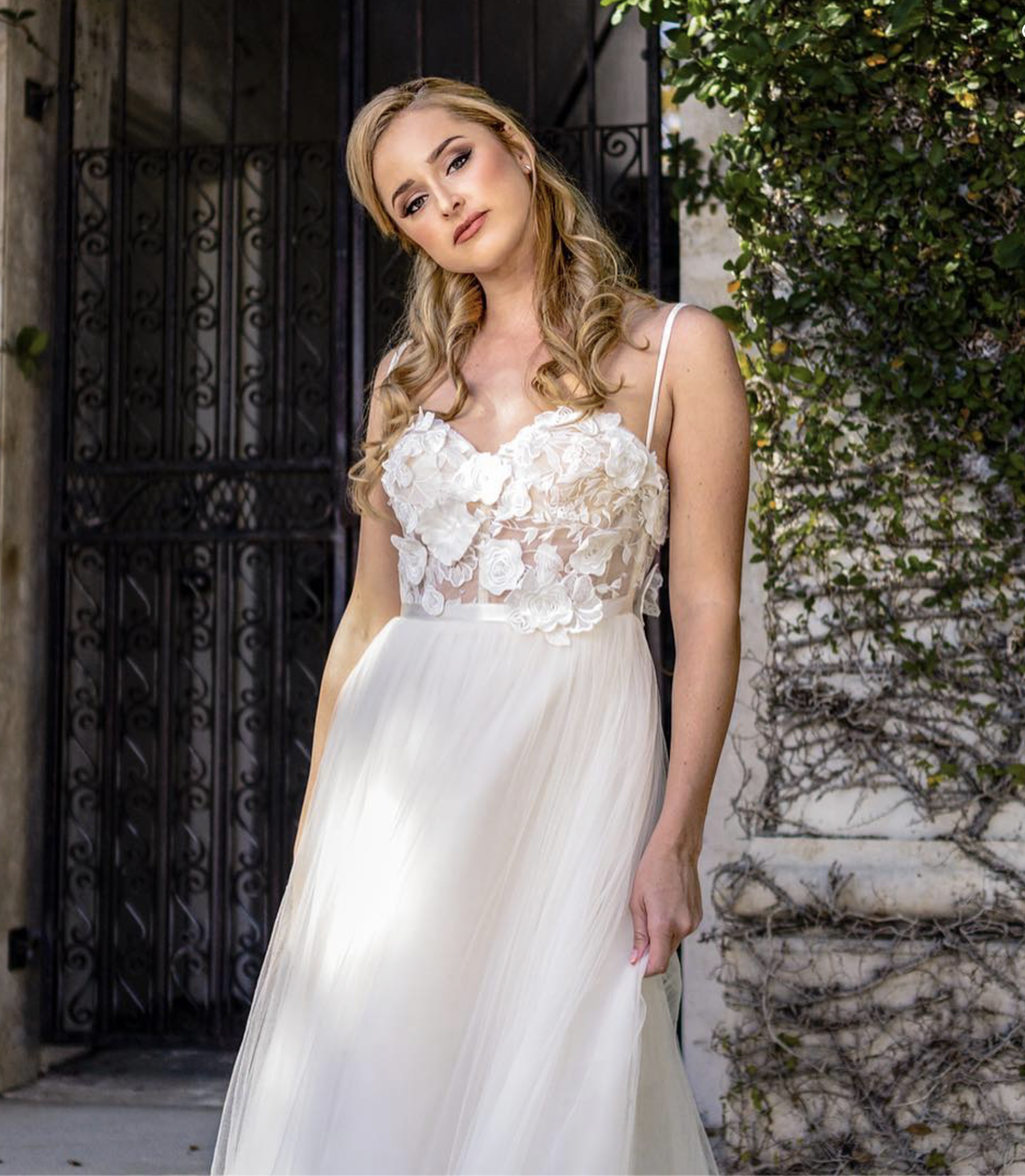 A young woman with blonde hair in loose curls wearing a white dress with floral appliqué on the bodice and thin straps, standing outdoors with a decorative black metal gate and green vine-covered wall behind her.
