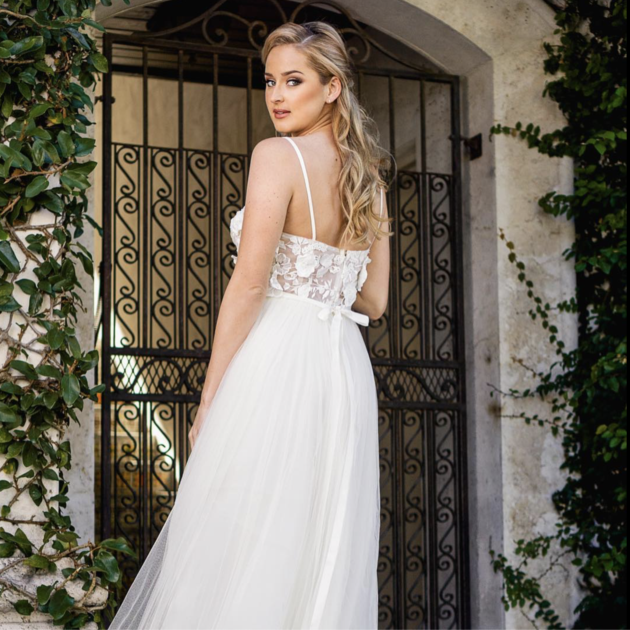 A woman in a white dress with lace details on the top, standing outdoors near a wrought iron gate and stone walls, surrounded by greenery.