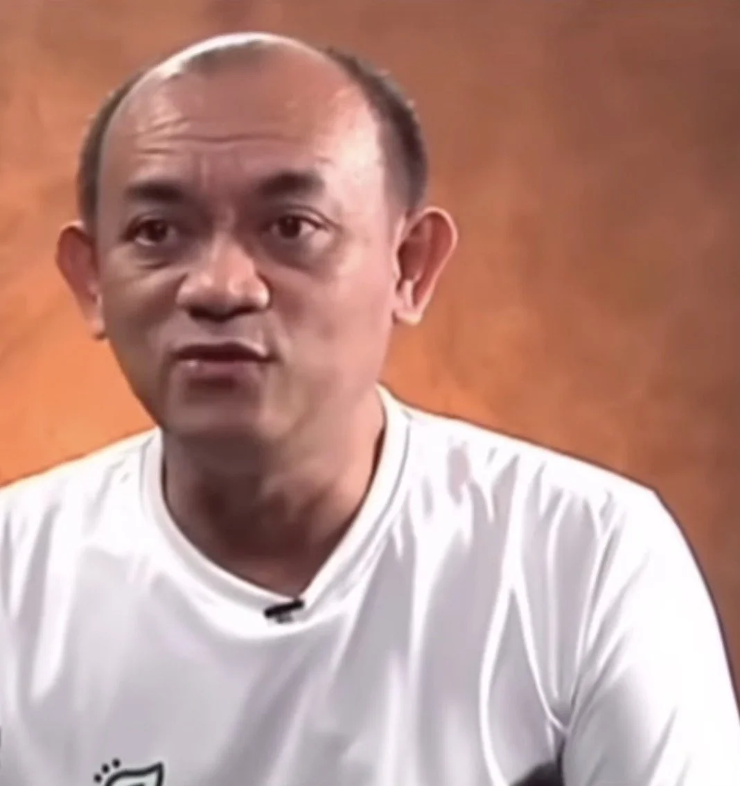 A man with a bald head and wearing a white shirt, speaking in front of a warm, orange-brown background.