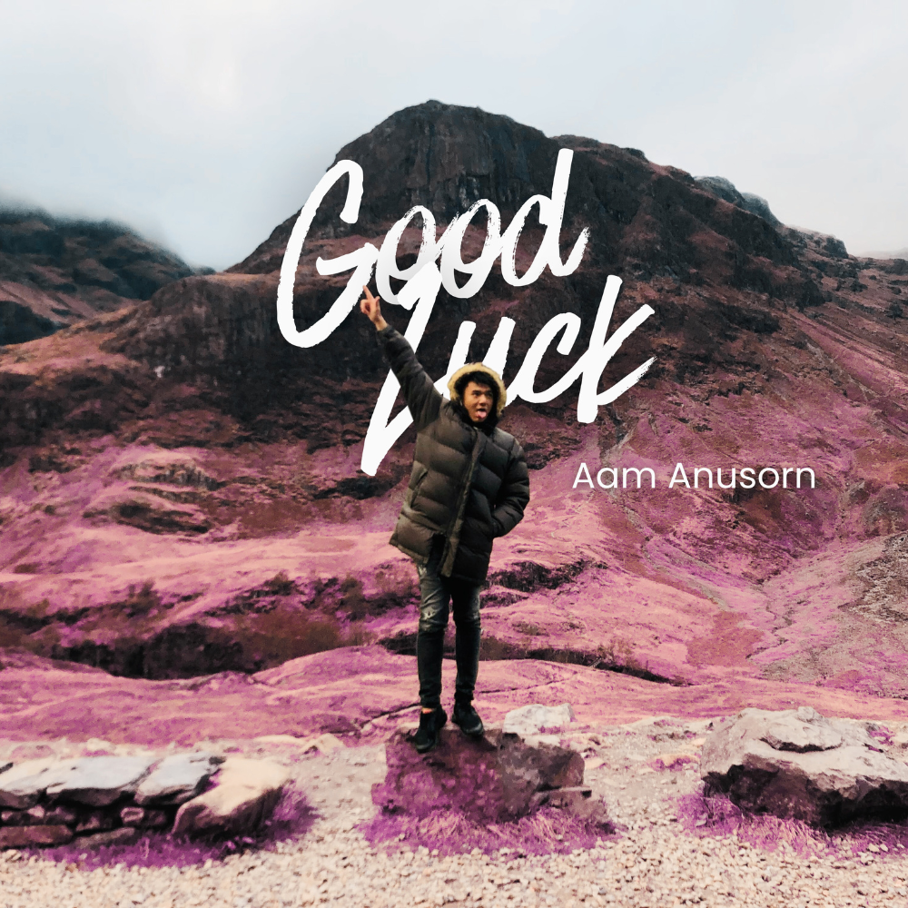 Person in a black jacket standing on a rock in a mountainous landscape with purple and brown hues, pointing upwards, with the text "Good Luck" and "Aam Anusorn" overlaid.