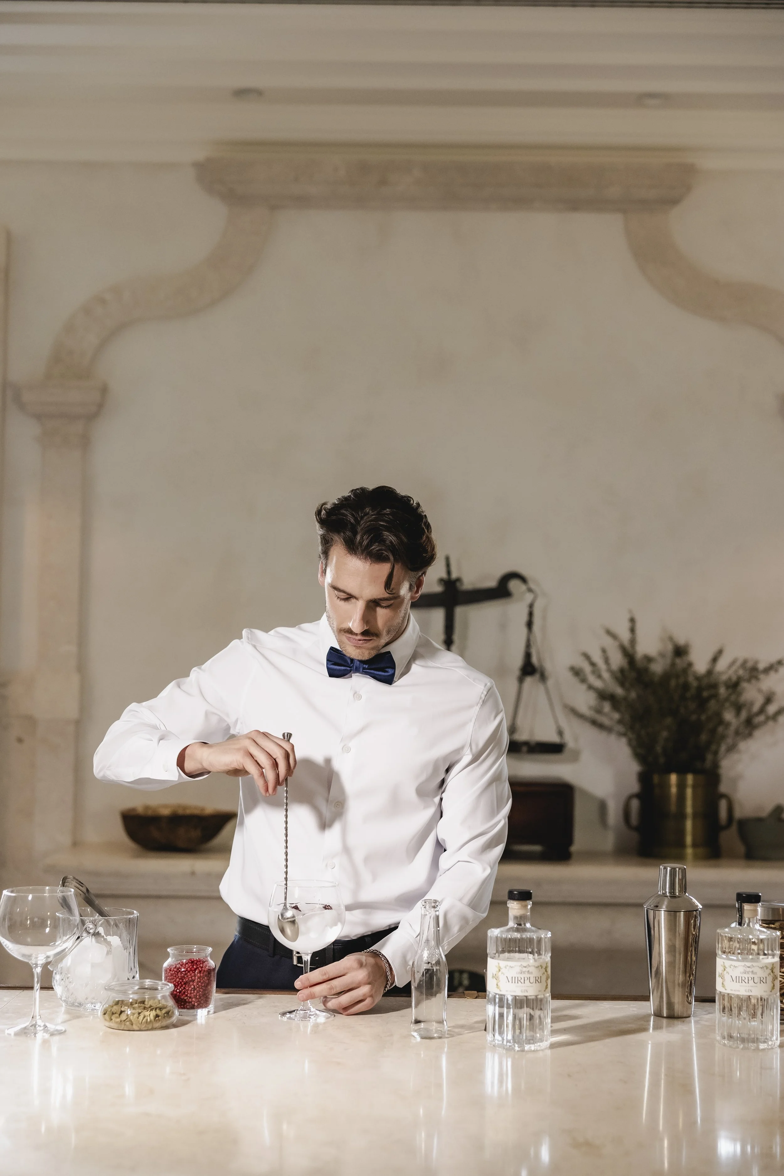 A man in a white shirt and blue bow tie preparing a cocktail in a large glass, surrounded by bar tools and ingredients on a marble countertop in a sophisticated, classic-style bar with a scale and potted plant in the background.