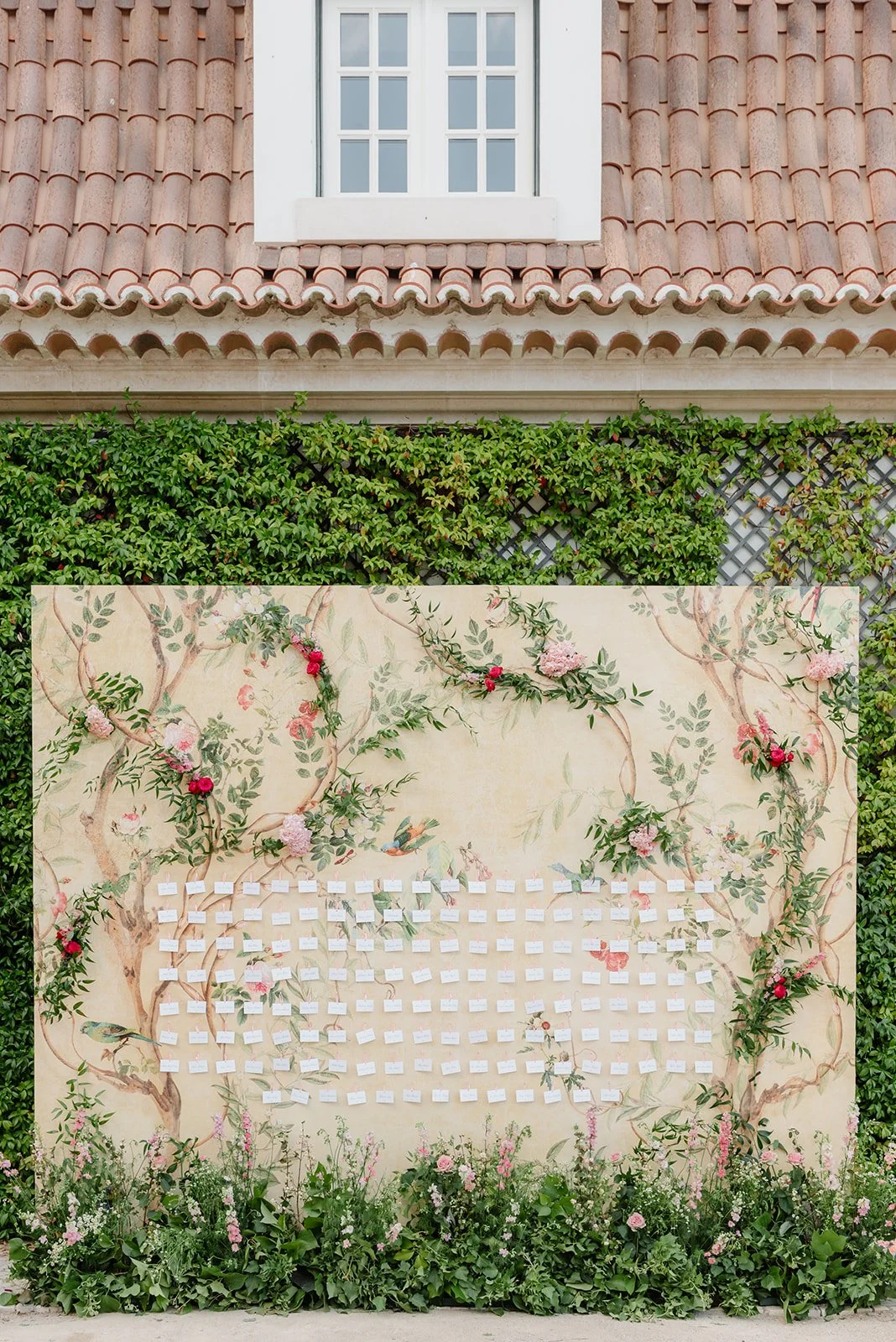 A large decorative display with a nature-themed mural featuring vines, flowers, and a bird. The display holds numerous small white cards pinned to it, arranged in a grid pattern. There are pink and red flowers at the bottom and greenery surrounding t