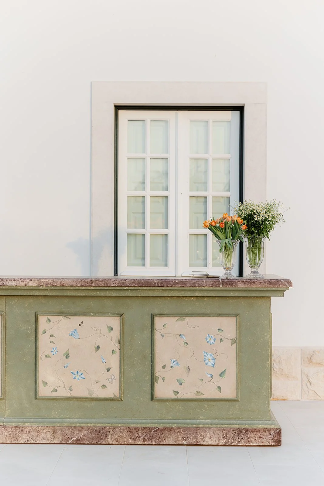 A white-framed window with white shutters above a decorative green counter with floral and bird designs, topped with two vases of flowers: one with orange tulips and the other with white baby's breath.
