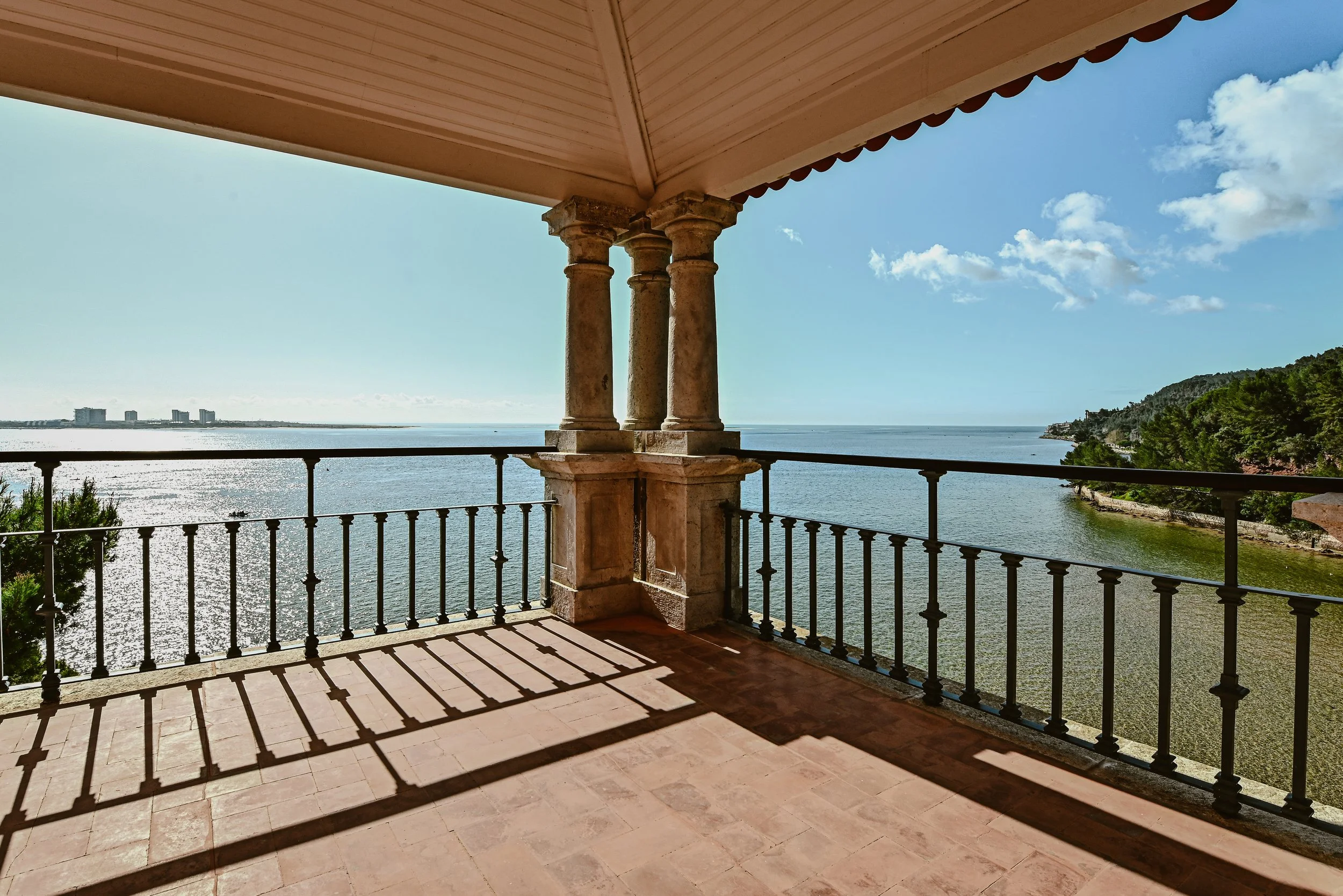 View from a balcony with stone columns overlooking a body of water, with hills and buildings in the distance, under a partly cloudy sky.