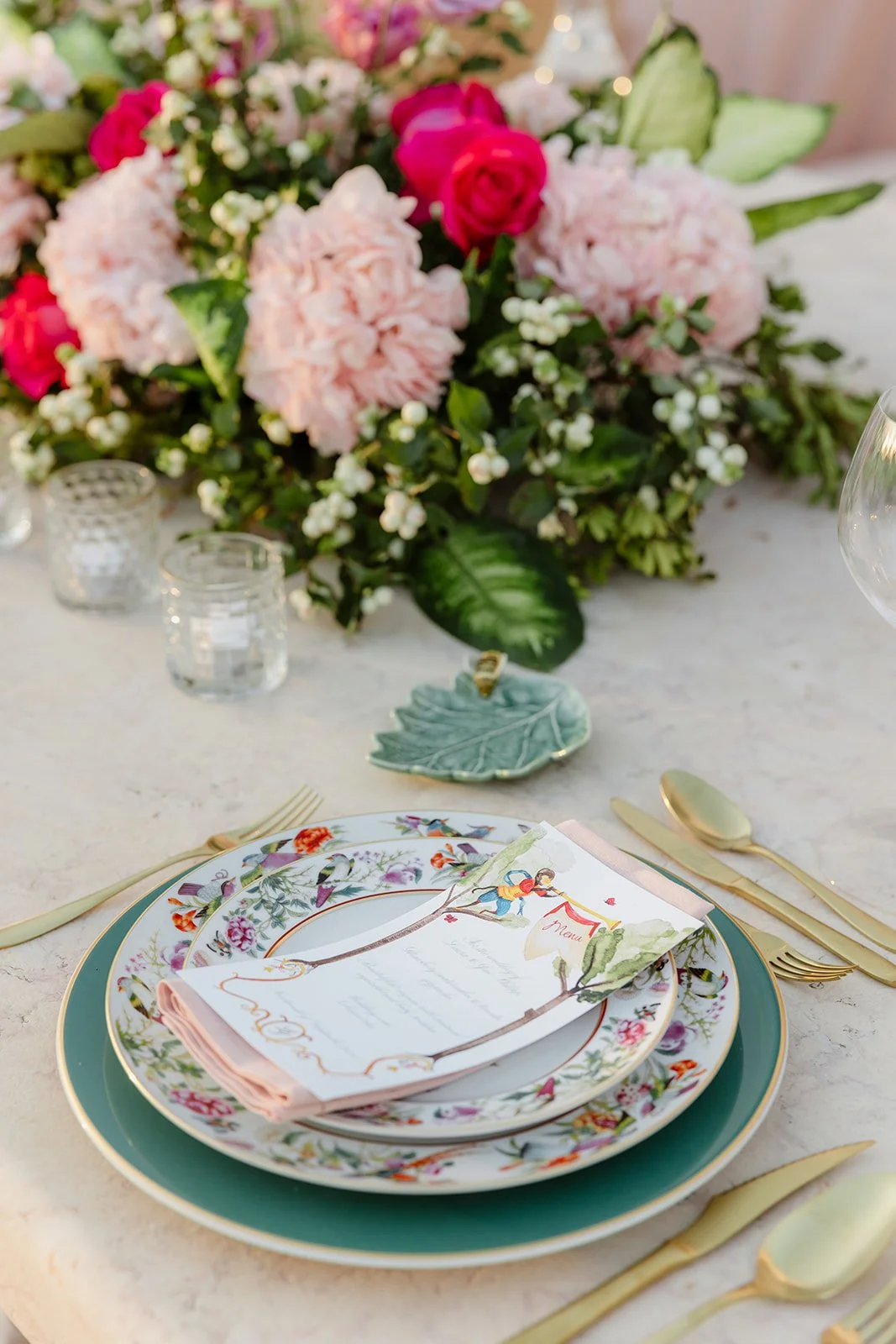 A table setting with floral patterned plates, gold utensils, a menu card, and a large bouquet of pink and red flowers with green leaves in the background.