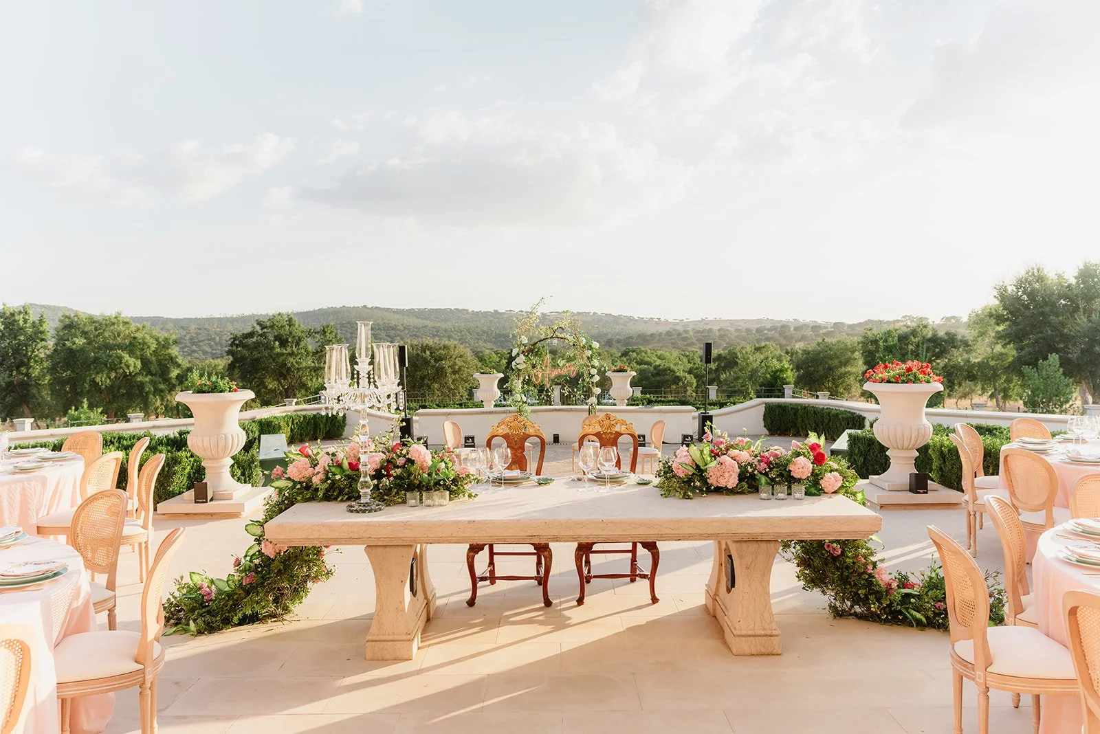 Elegant outdoor wedding reception setup with a decorated table, floral arrangements, chairs, and scenic view of trees and hills in the background.