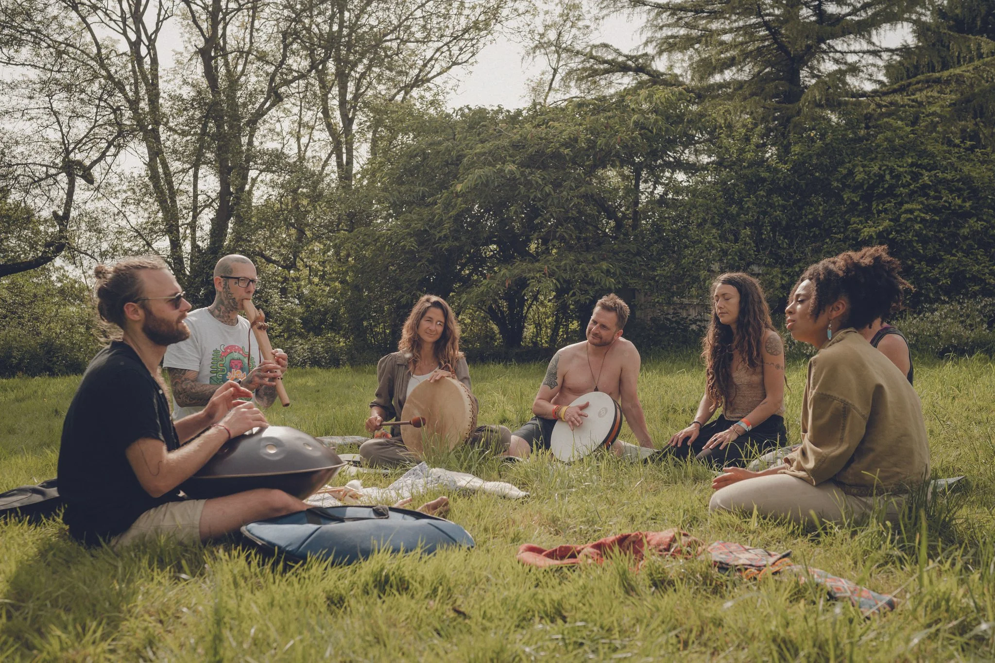 Group of seven people sitting on the grass in a park, playing musical instruments and meditating surrounded by trees.