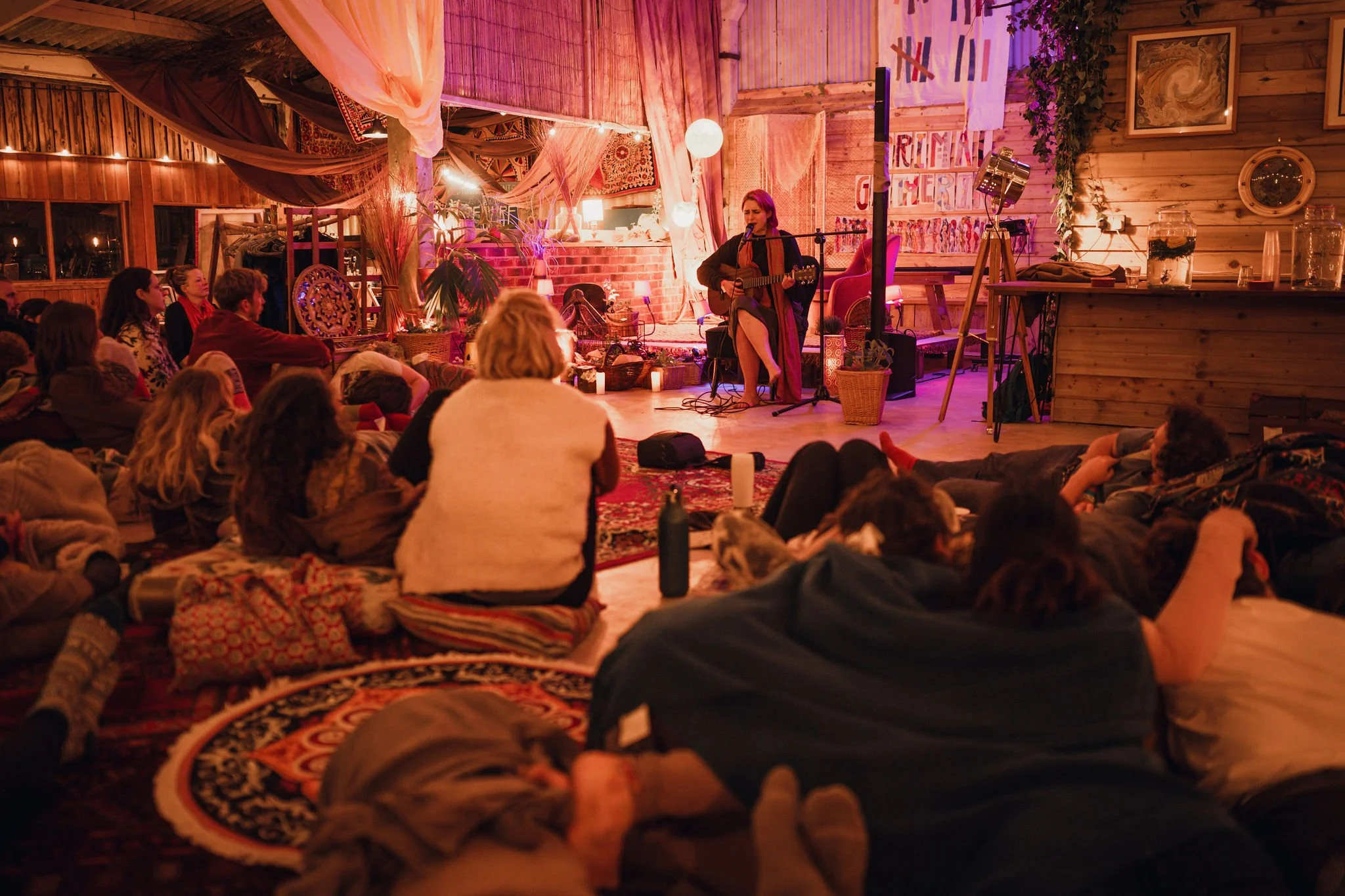 A woman playing guitar and singing on a small stage at a cozy indoor venue decorated with warm lighting, plants, and colorful textiles, with an audience seated on the floor and cushions watching her perform.