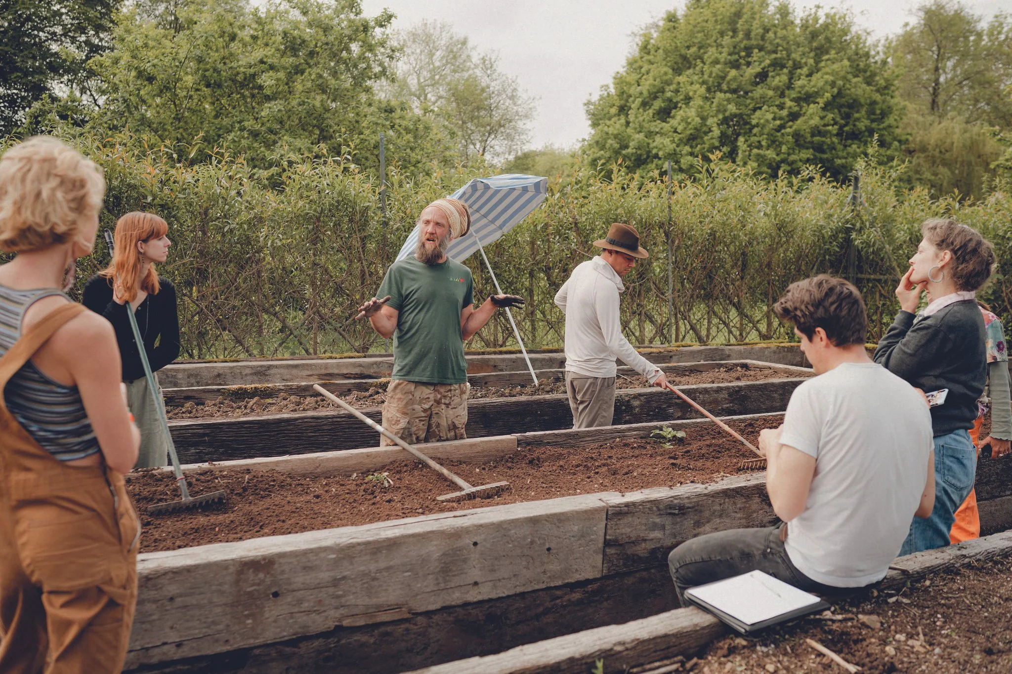 Group of people in a garden or farm discussing gardening or farming techniques, with some holding gardening tools and a person speaking to the group.
