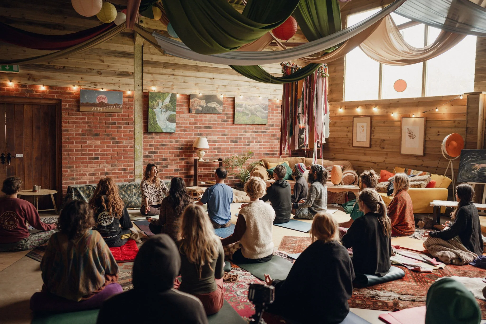 A group of people sitting on yoga mats in a cozy, wooden room with brick walls, attending a meditation or yoga session led by an instructor sitting on a cushion at the front. The room is decorated with colorful fabric drapes, framed artwork, and stri