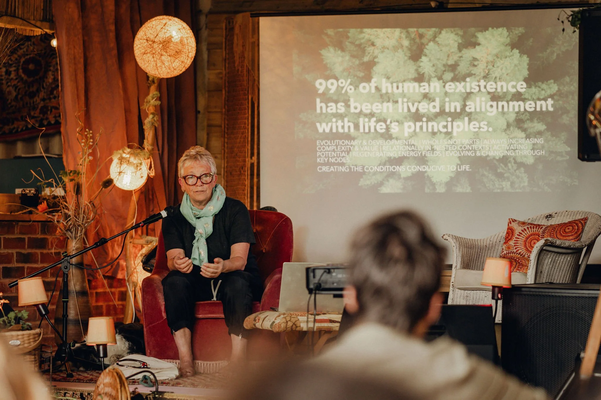 An older woman with glasses and a mint green scarf sitting on a red armchair gives a presentation in a warmly decorated room. A slide projected on the wall behind her reads '99% of human existence has been lived in alignment with life's principles.'