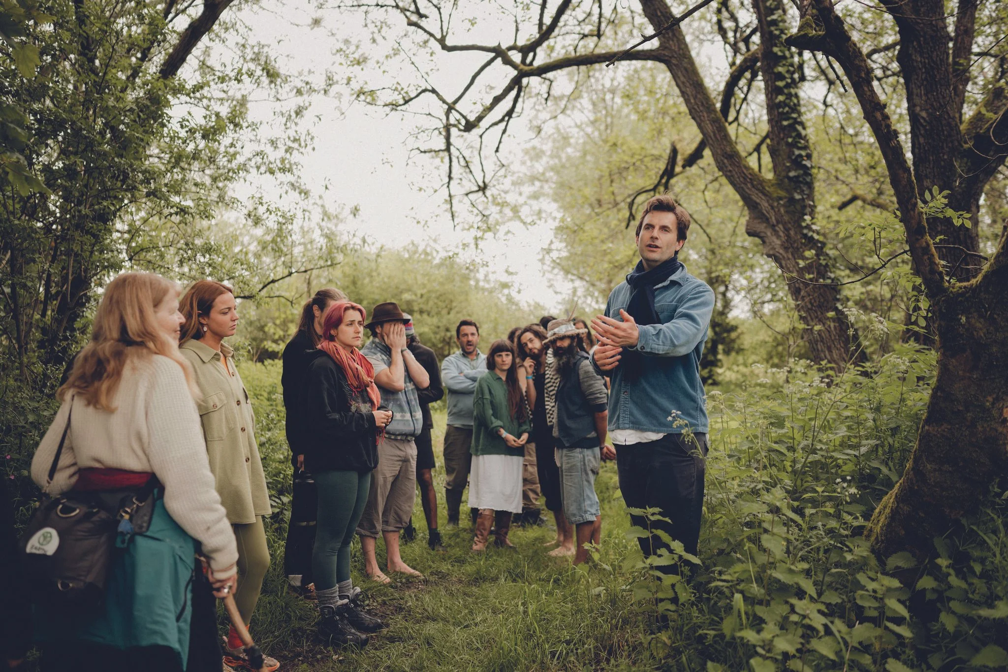 A group of people gathered outdoors in a wooded area, listening to a man in front of them, who appears to be giving a tour or lecture. The group includes men and women dressed casually, some with backpacks, standing amidst trees and greenery.