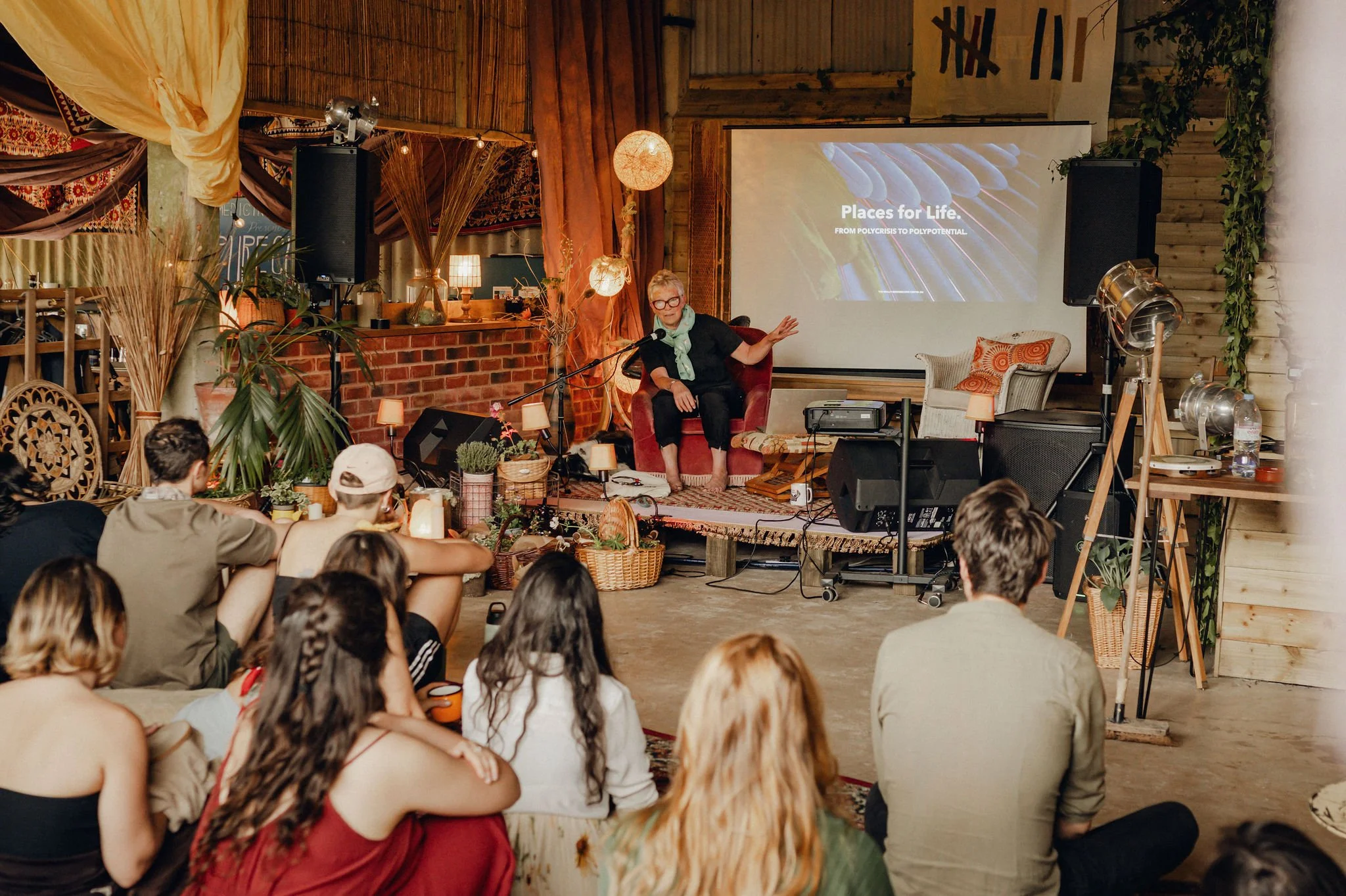 A woman giving a presentation or lecture on a small stage decorated with plants and warm lighting to an audience sitting on the floor and chairs in a cozy, rustic venue.