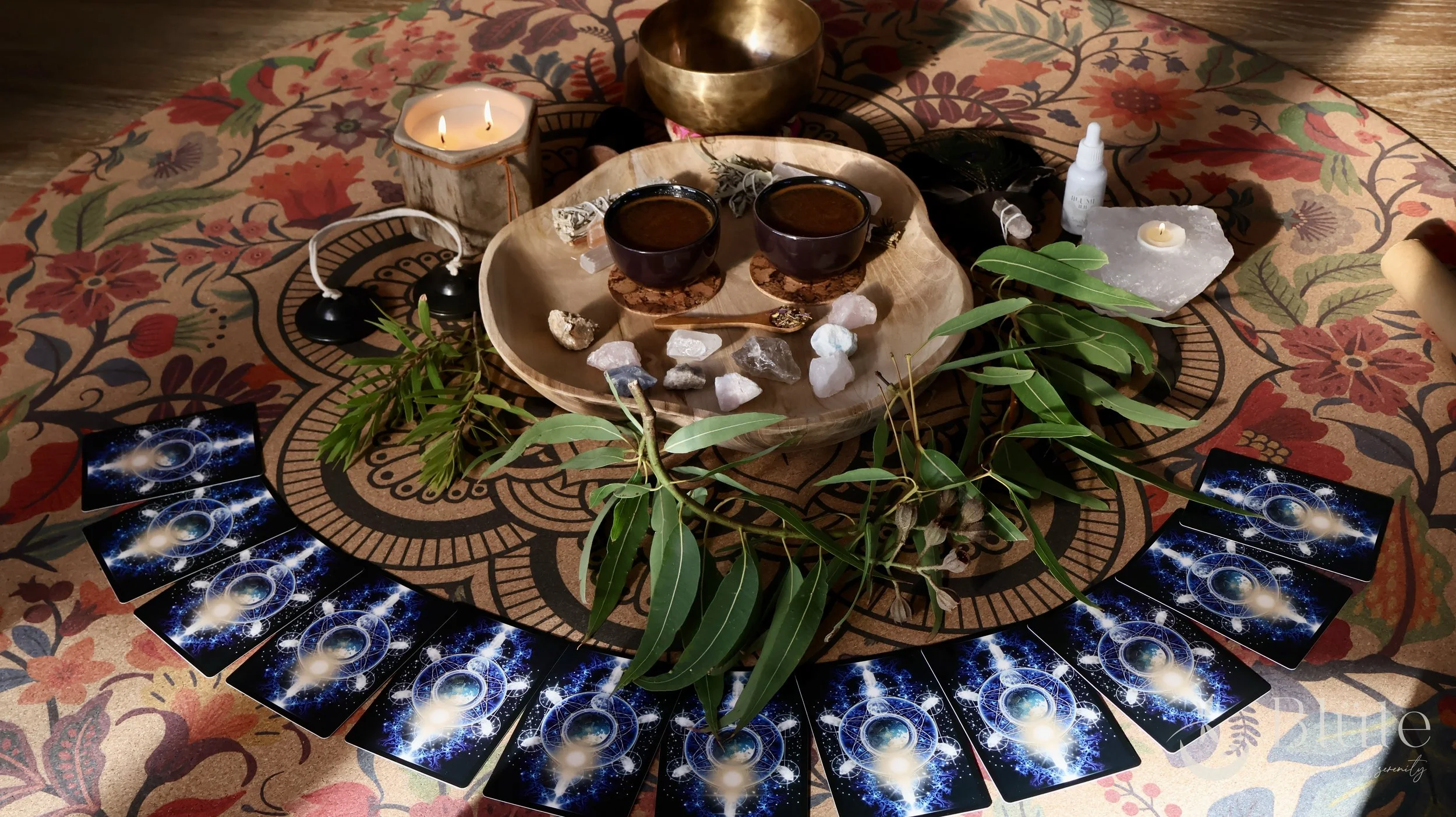 A round table with a colorful floral tablecloth holding a wooden tray with two cups of tea, crystals, rocks, and small bottles, surrounded by green leaves and tarot cards arranged in a semi-circle, with candles and a singing bowl in the background.