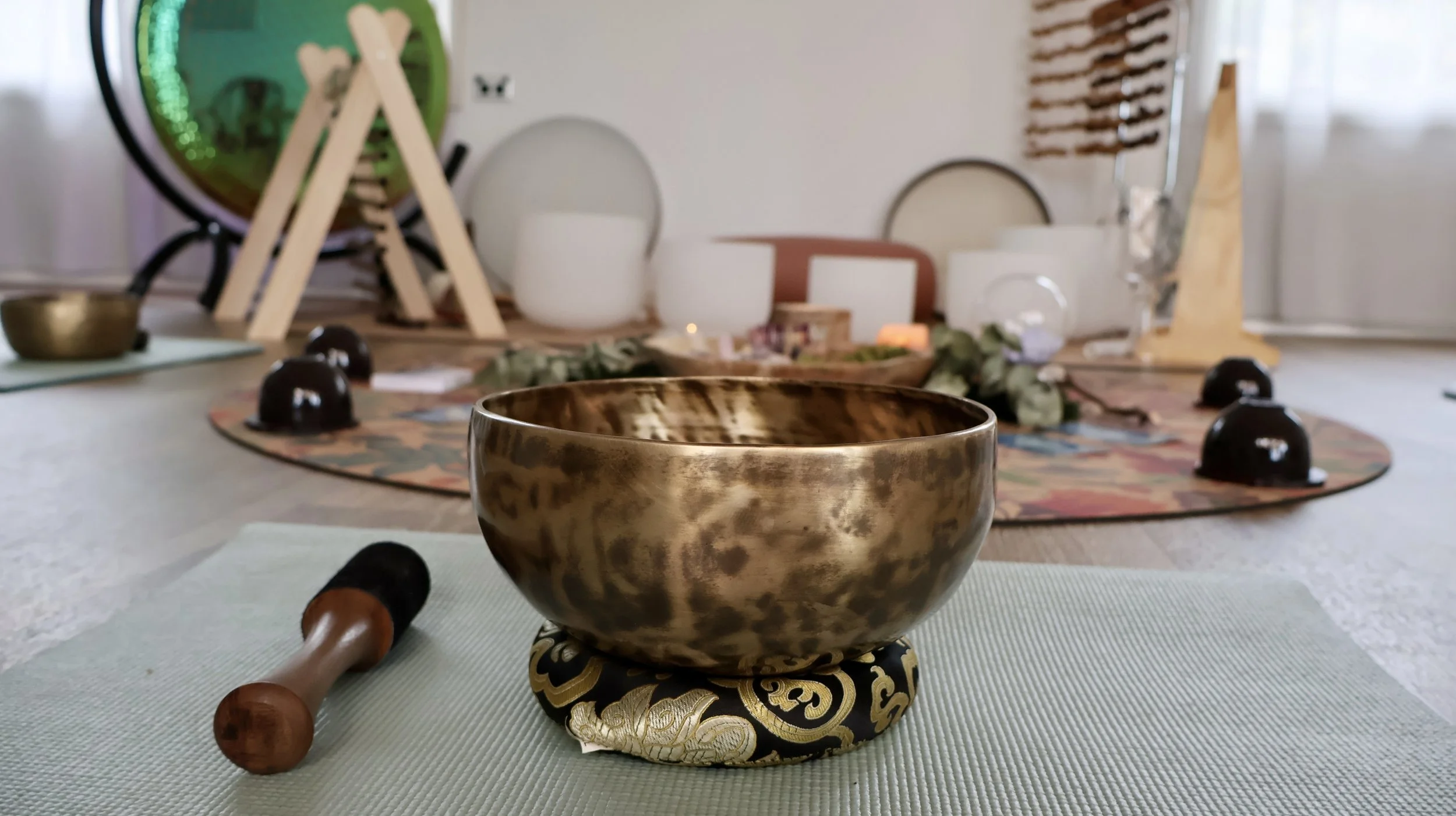 A singing bowl made of polished metal with a wooden mallet lying beside it, on a textured mat in a room set up for meditation or sound healing with various instruments and decorative items in the background.