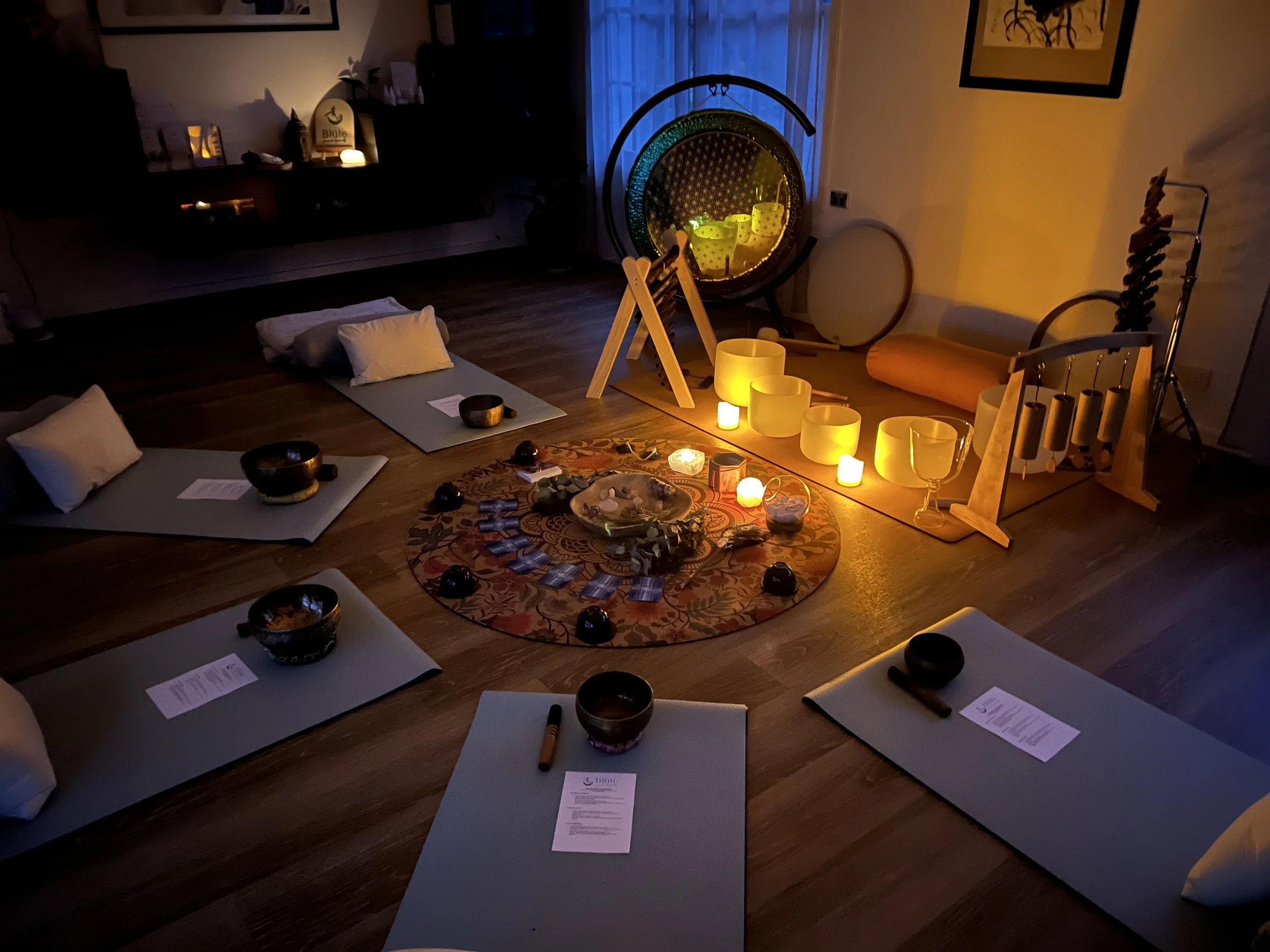 A meditation space set up indoors with five yoga mats arranged in a circle, each with a pillow and a bowl. In the center, there is a circular carpet with crystals and candles. Various singing bowls and chimes are placed around the space, with soft candle and ambient lighting creating a warm, calming atmosphere.