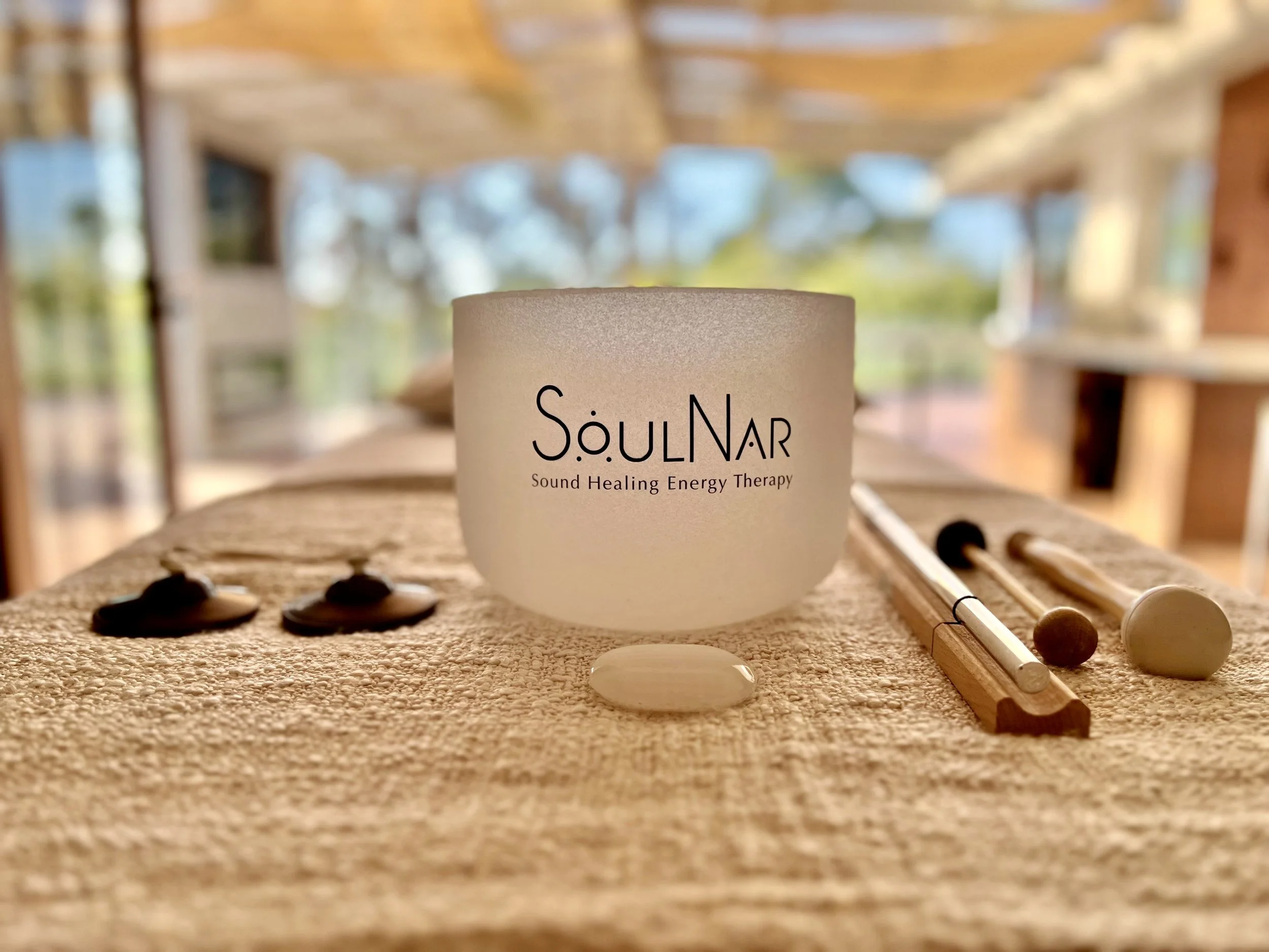 A sound healing bowl labeled "SoulNAr Sound Healing Energy Therapy" is placed on a textured surface with three smaller objects and two wooden mallets nearby, inside a bright room with large windows showing trees outside.