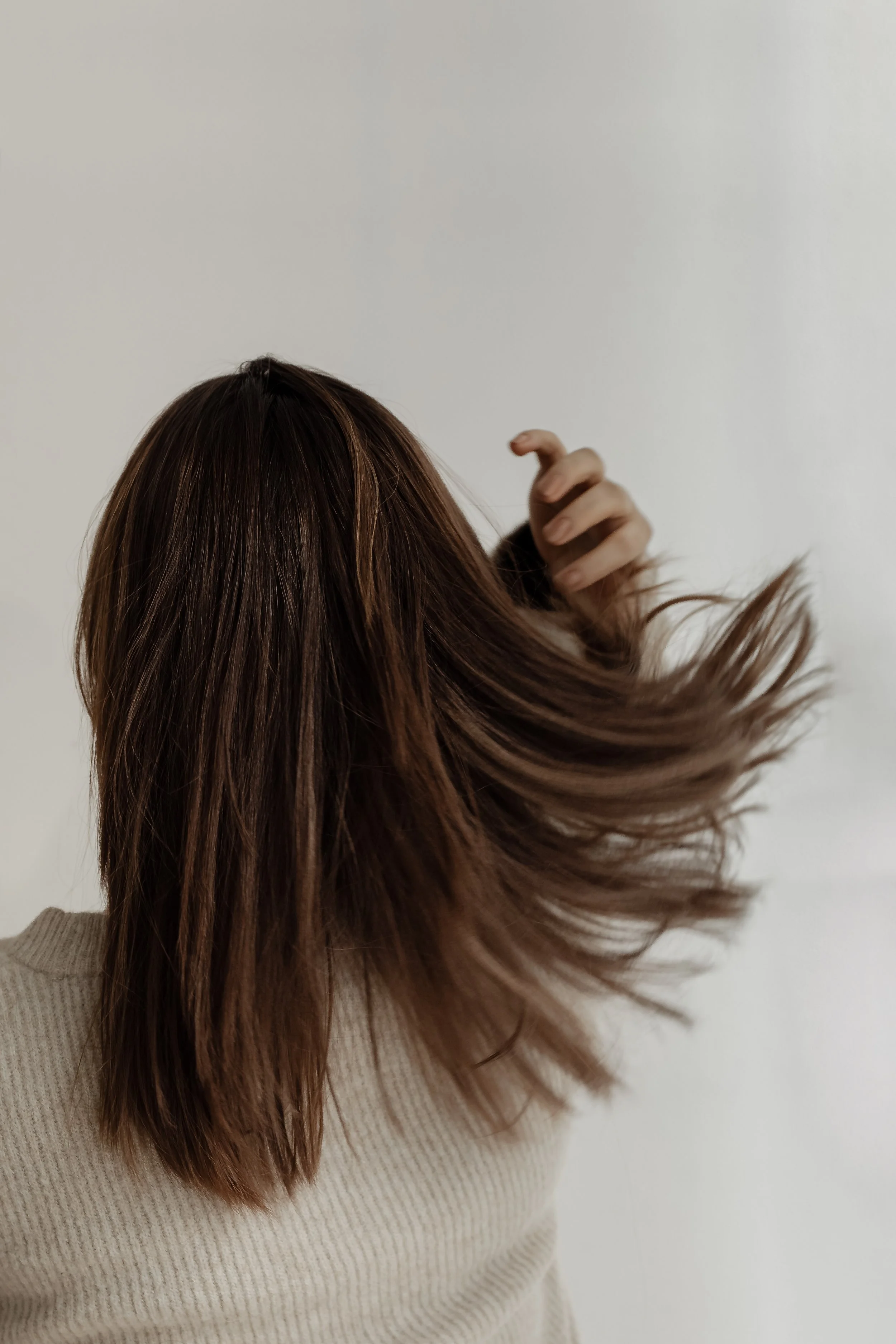 Woman with brown hair flipping her hair, wearing a beige sweater, against a plain white background.