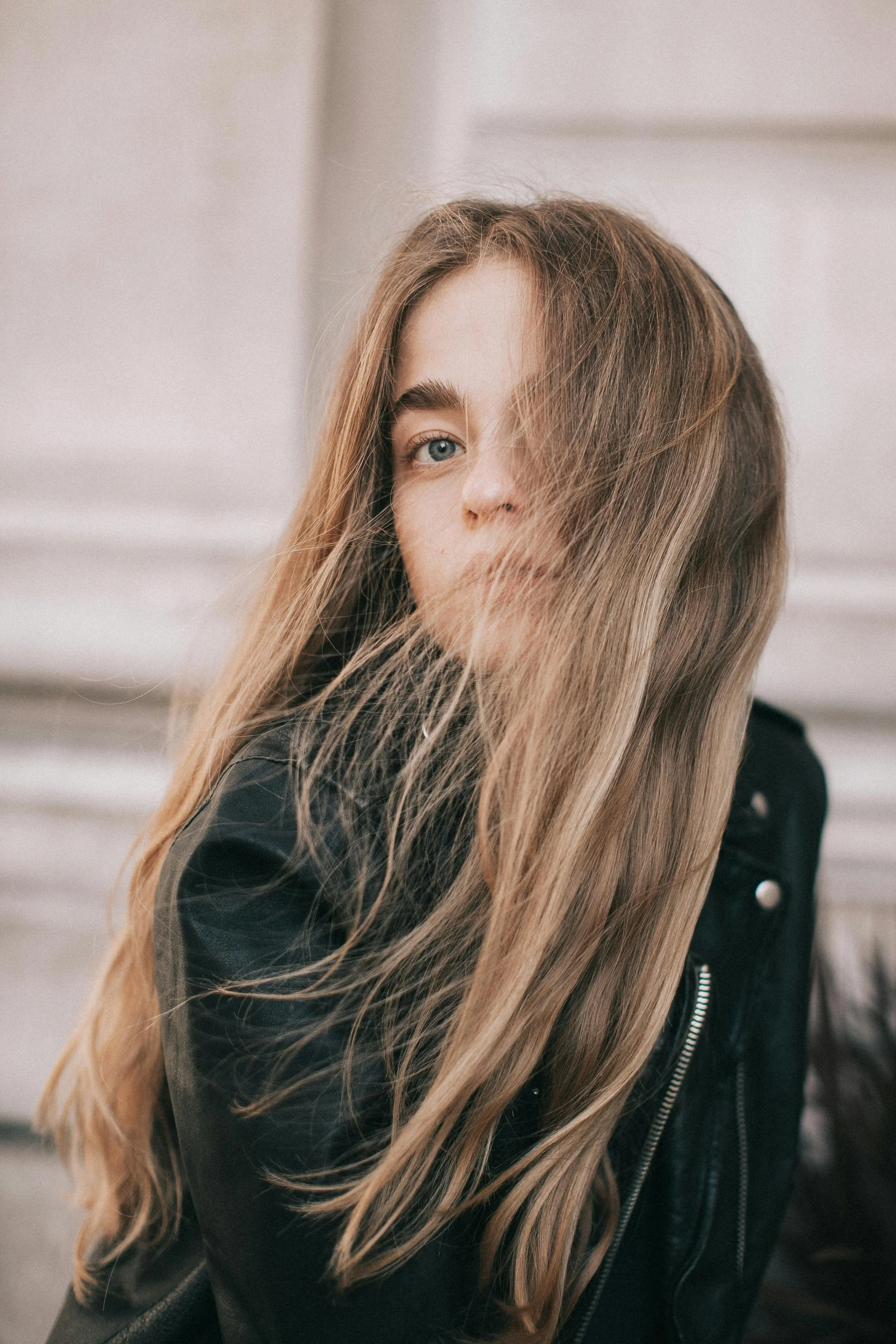 A young woman with long, wavy blonde hair partially covering her face, wearing a black leather jacket, in an indoor setting.