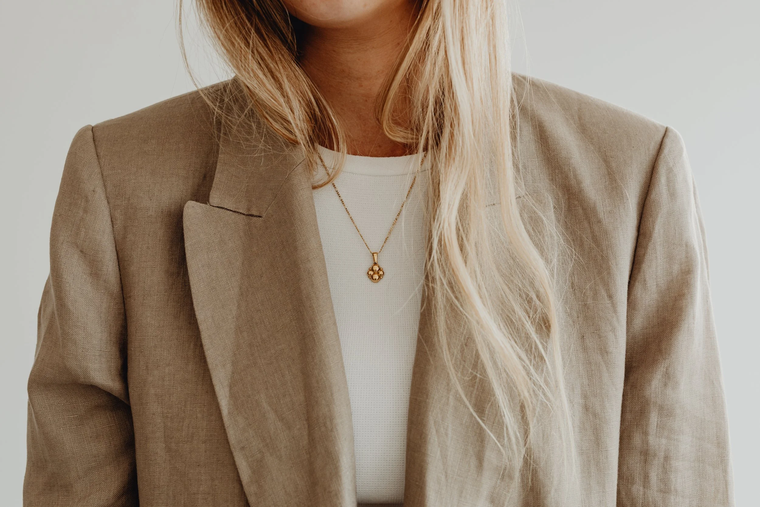 Close-up of a person's torso wearing a beige blazer, white shirt, and a gold necklace with a pendant, with blonde hair over the shoulders.