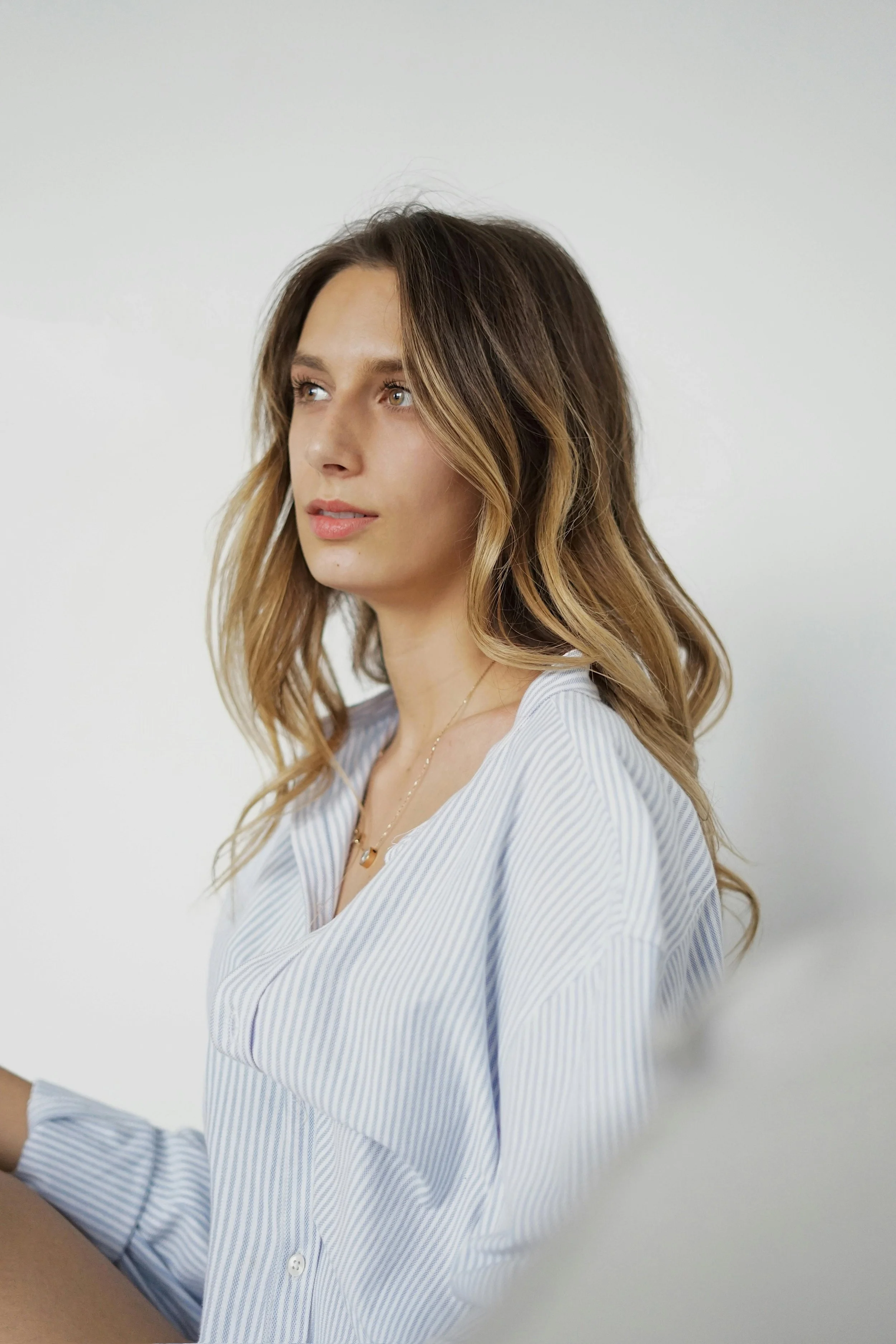 A young woman with long, wavy, light brown hair wearing a blue and white striped blouse, looking to her left against a plain white background.