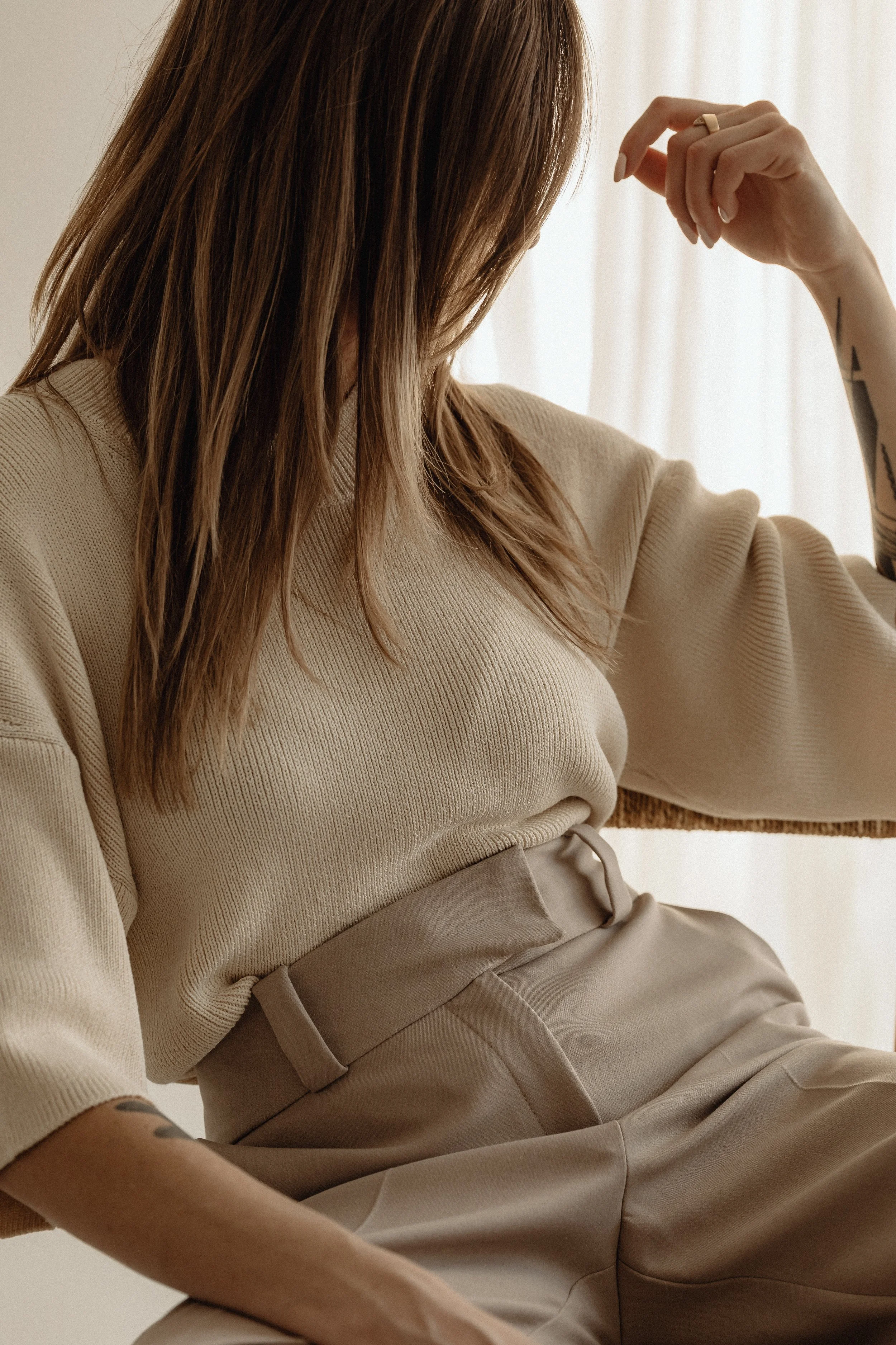 Woman with brown hair wearing beige sweater and beige pants, sitting with her head down and hand near her face, in front of a white curtain.