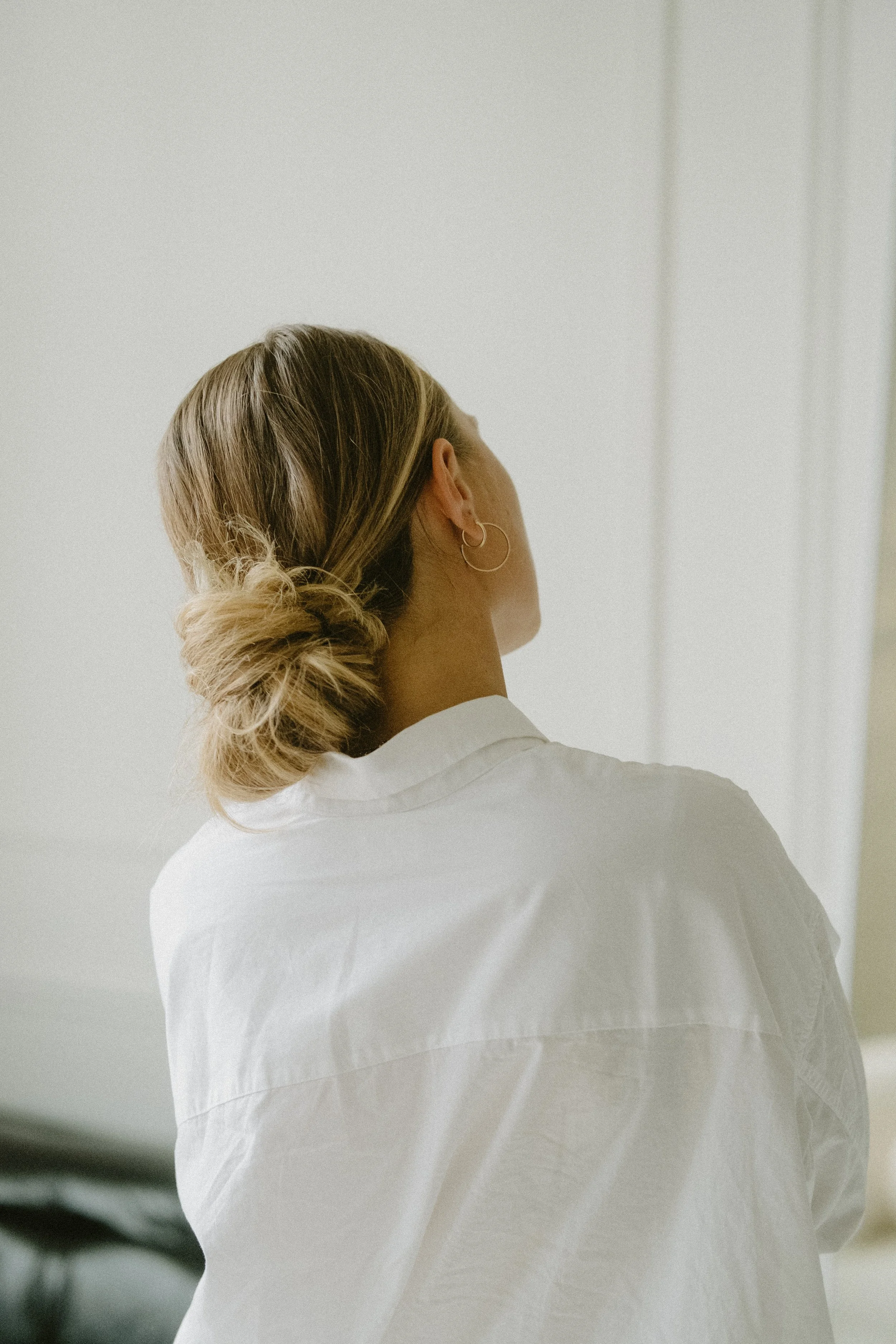 Back view of a woman with blonde hair styled in a loose bun, wearing a white shirt and hoop earrings, standing in a minimalistic room.
