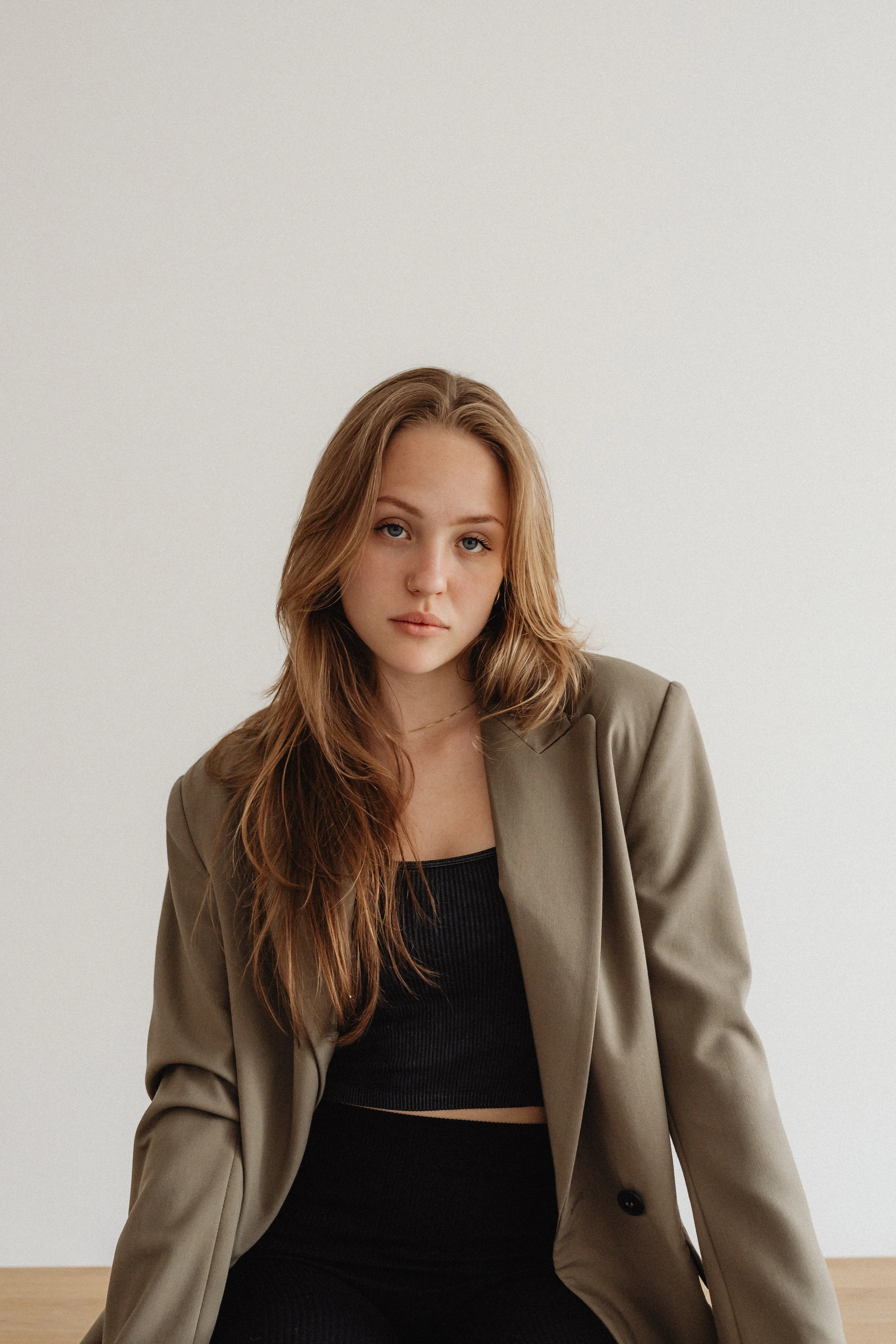 A young woman with long, wavy, reddish-brown hair and light skin, dressed in a black ribbed crop top and a beige blazer, sitting in front of a plain white background.