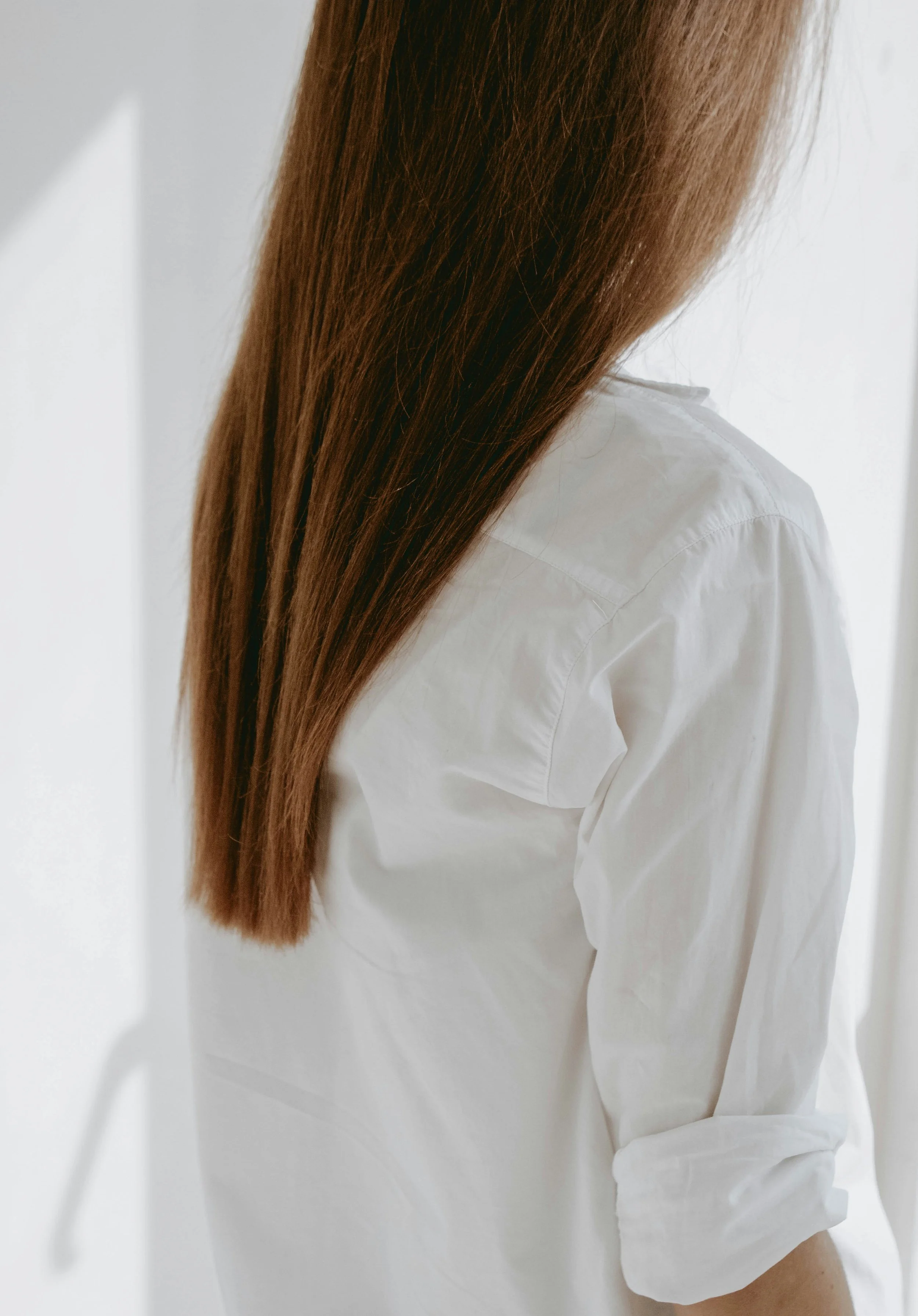 Close-up of a woman with long brown hair wearing a white shirt, standing near a white wall.