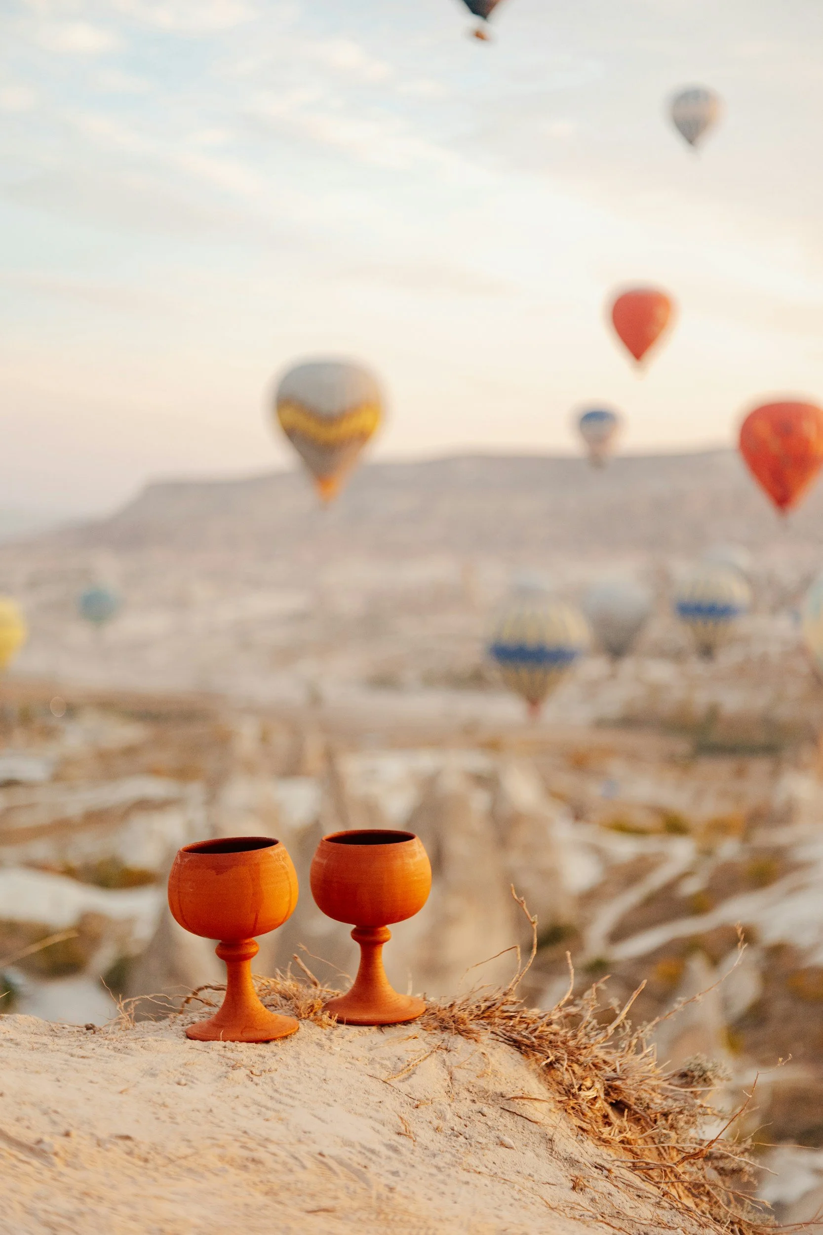 Two terracotta cups on a sandy surface with a blurred landscape of hot air balloons floating in the sky in the background.