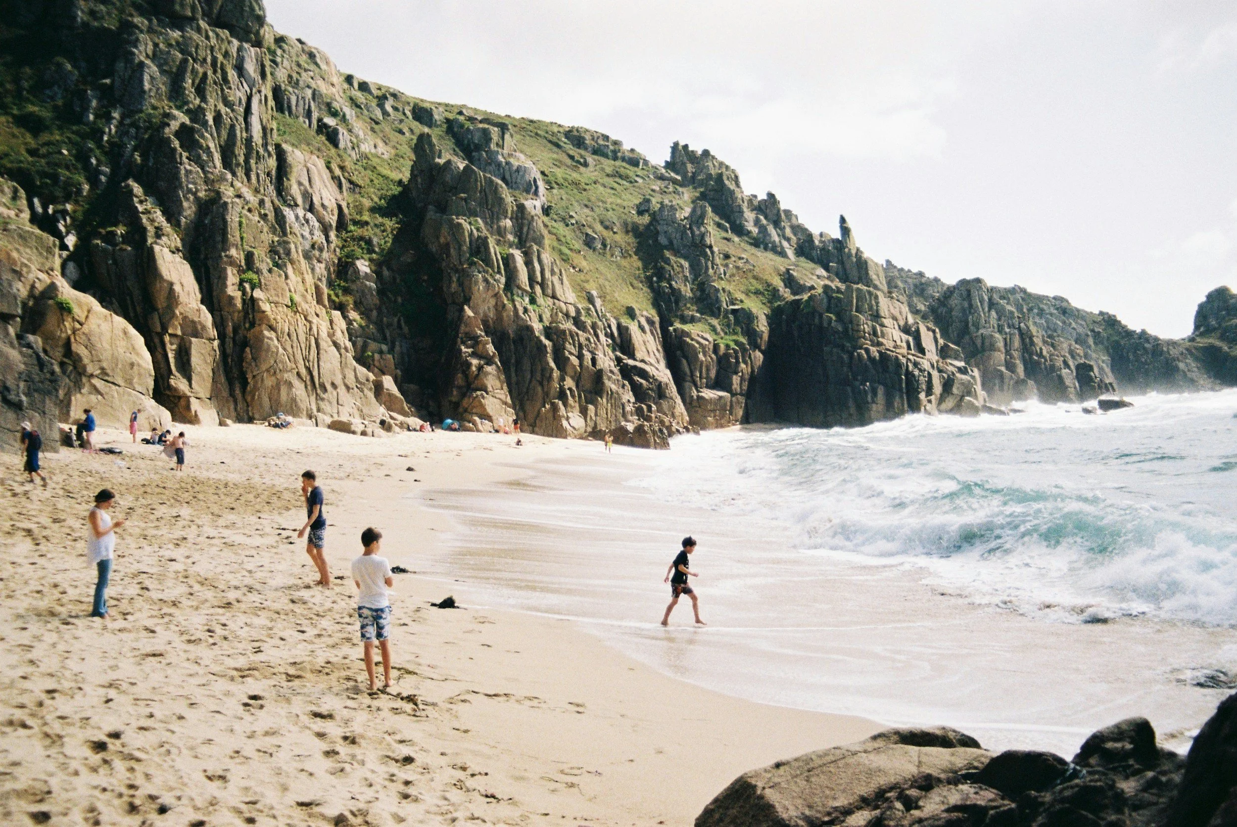 People standing and walking along a sandy beach with large rock formations and cliffs in the background, waves crashing on the shore.