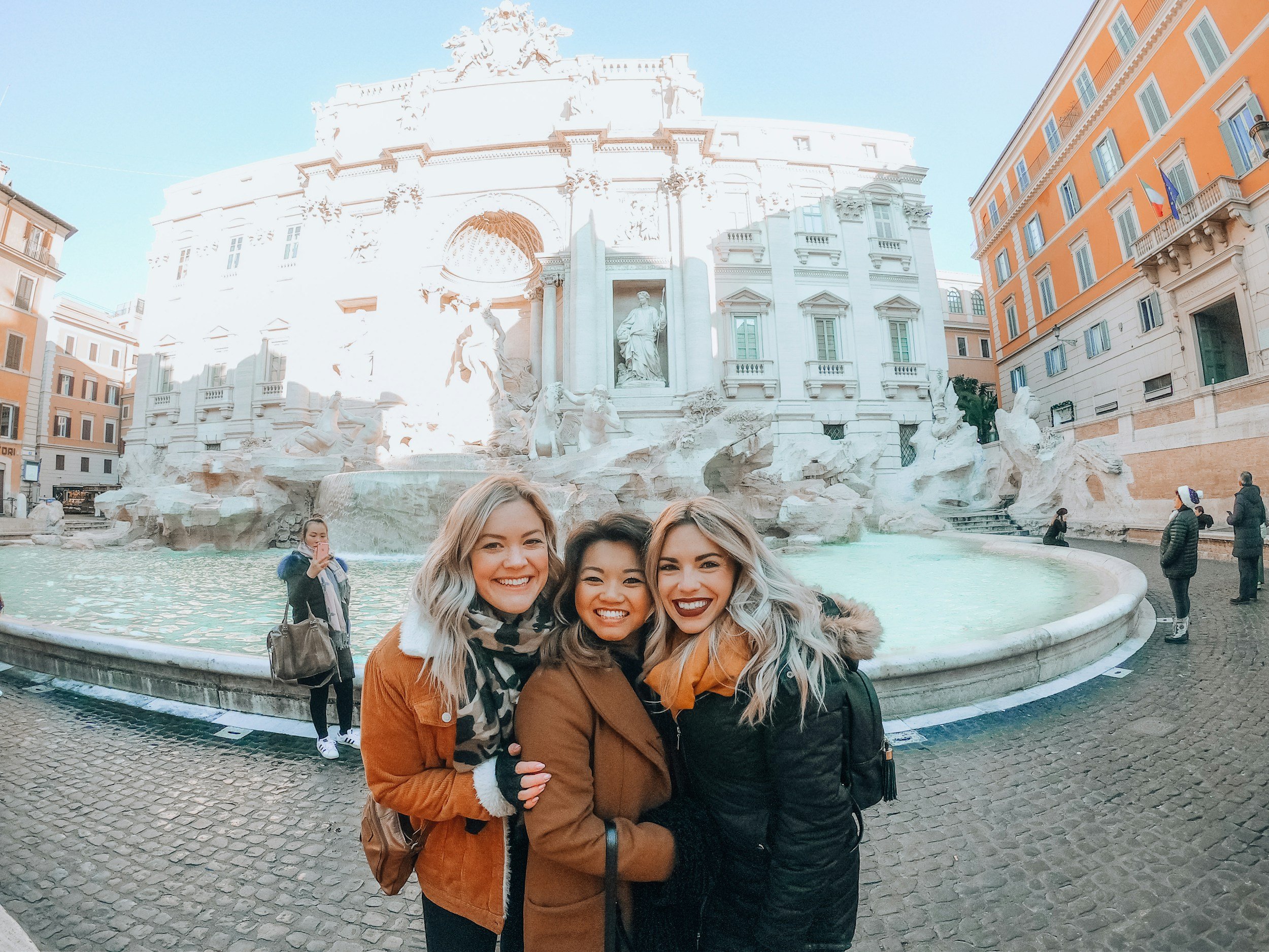 Three smiling women standing in front of the Trevi Fountain in Rome, Italy, with buildings and other visitors in the background.
