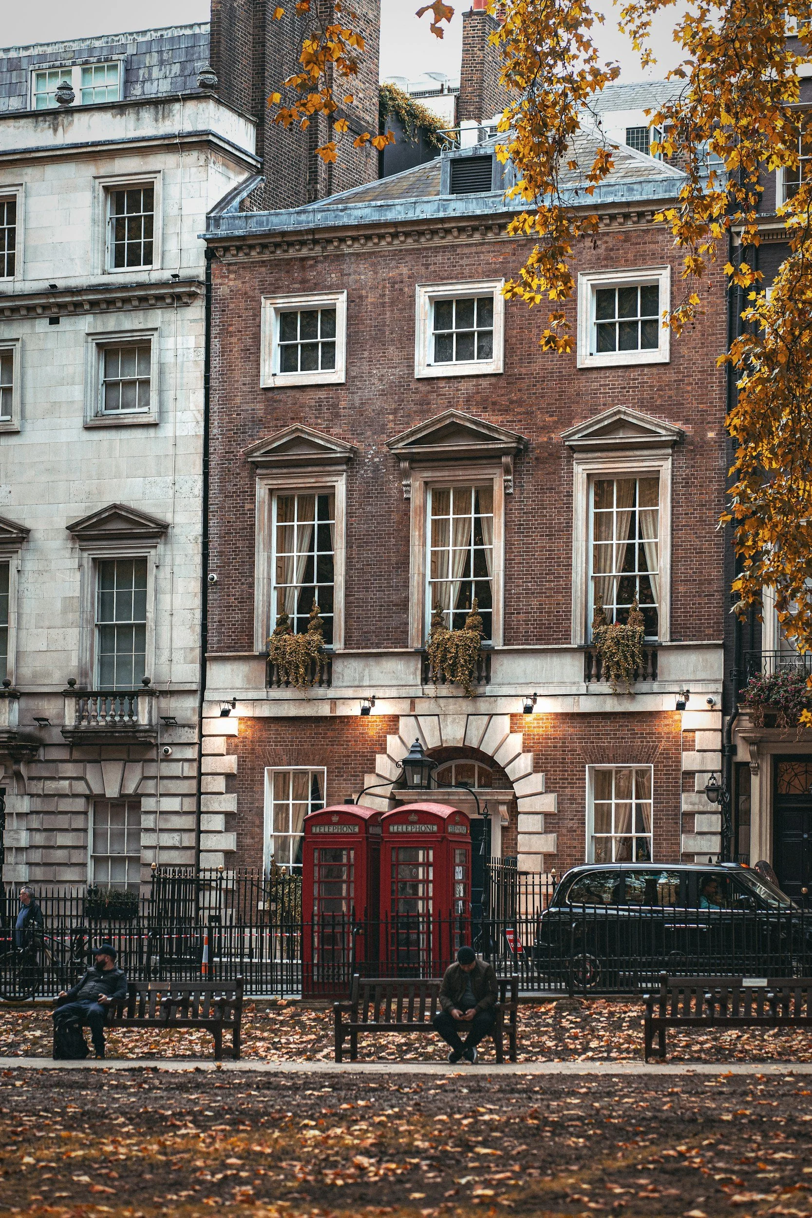 Street scene with old brick building, traditional red telephone booths, black taxi, pedestrians, autumn leaves, and benches.