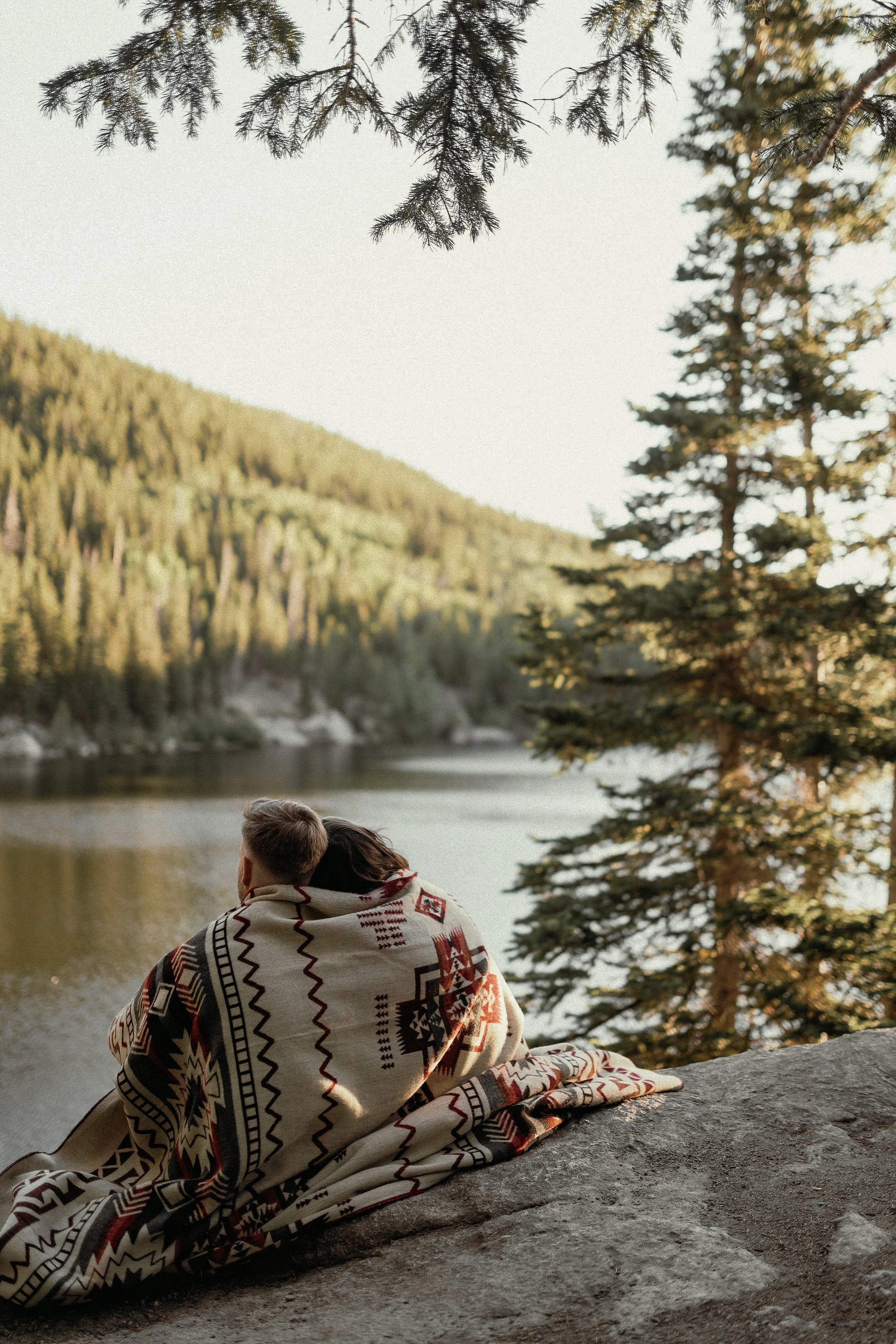 A couple sitting on a rock by a river, wrapped in a patterned blanket, surrounded by trees and mountains in a peaceful outdoor setting.