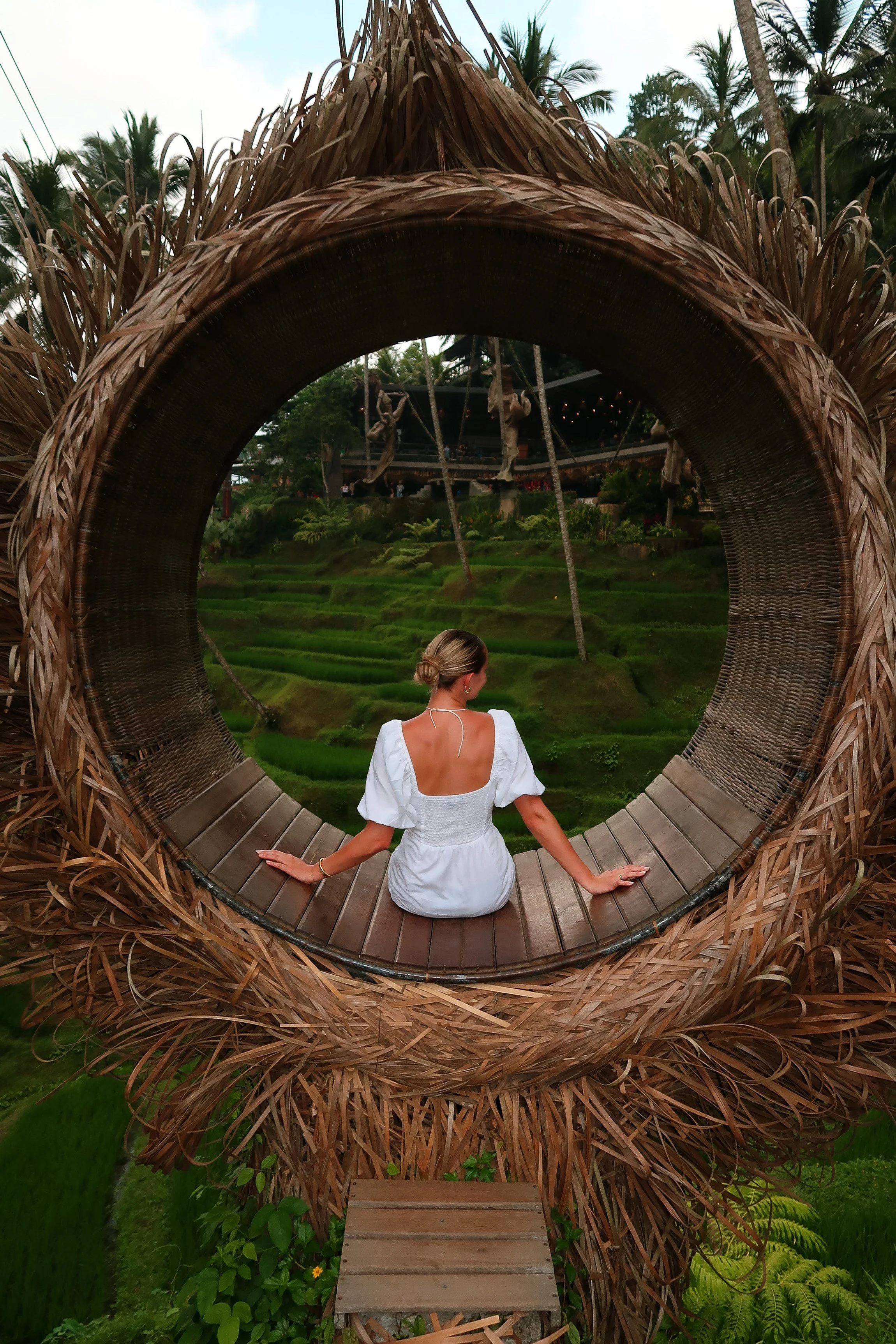 A woman in a white dress sitting inside a large, circular, woven bamboo structure overlooking lush green terraced rice paddies with palm trees and a building in the background.