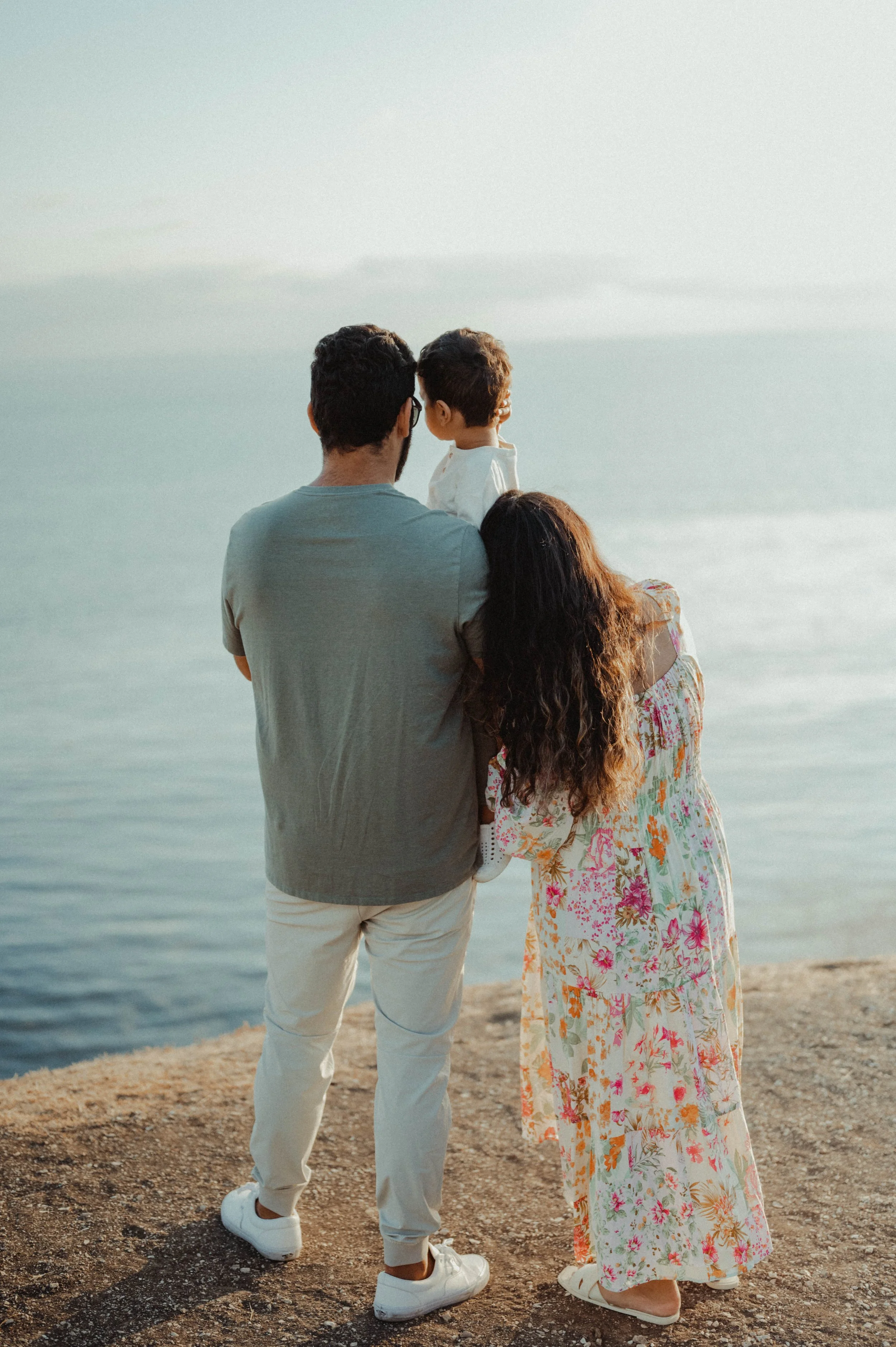 A family of three standing on a sandy shore, looking out at the ocean during sunset. The father holds a young boy, and the mother stands close, all facing away from the camera.