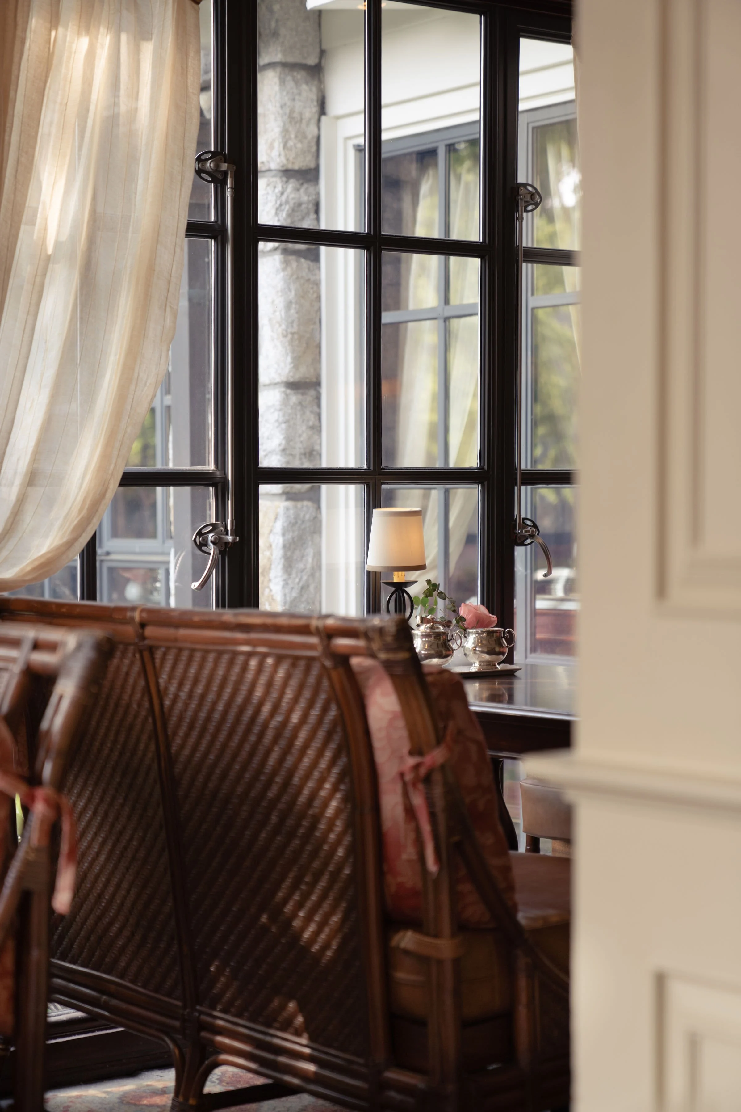 A cozy corner with black-framed windows letting in natural light, a small table with a lamp, teapots, and a plant, and part of a rattan chair with floral cushions.