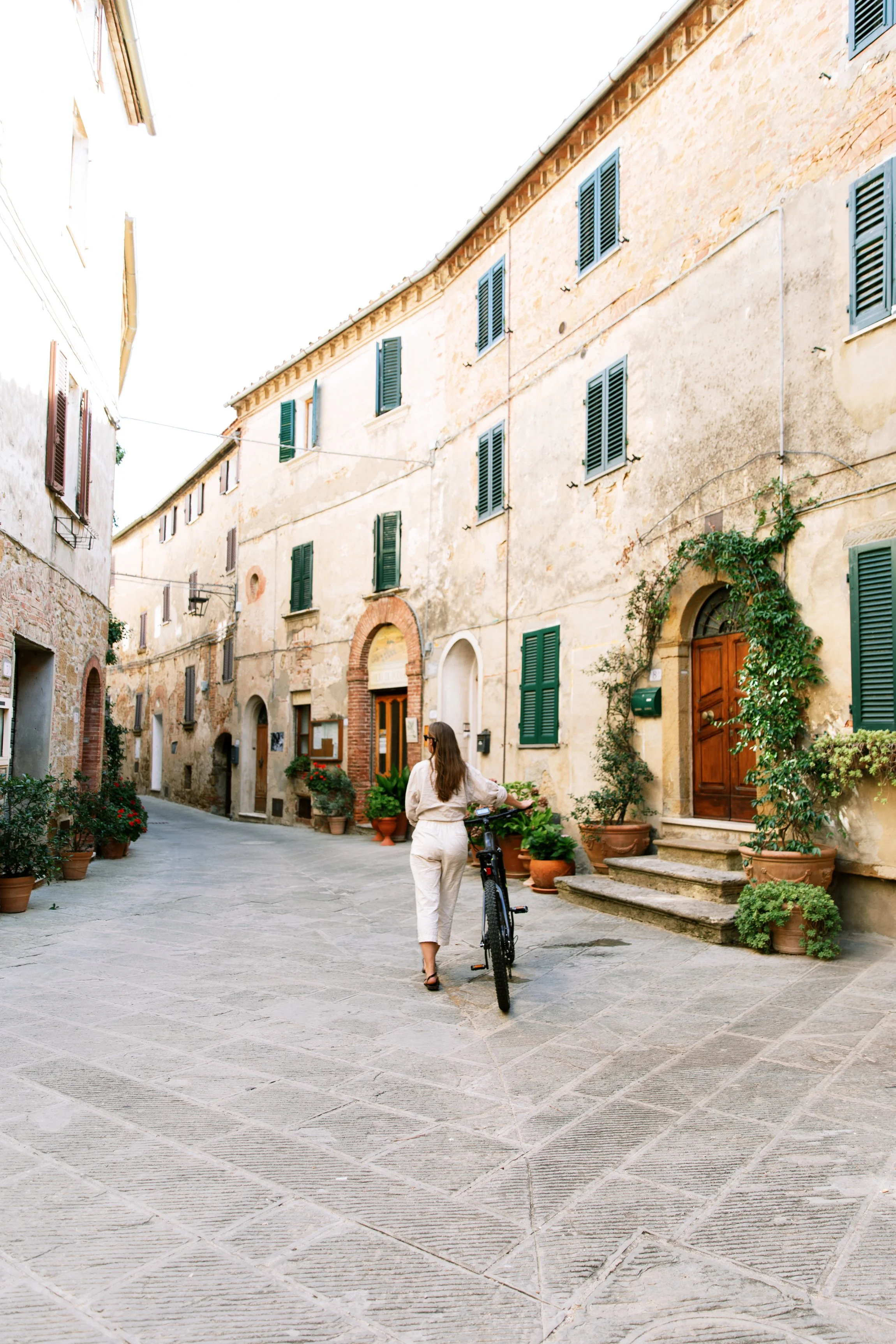 A woman walking her bicycle down a narrow European street with old stone buildings, green shutters, and potted plants.