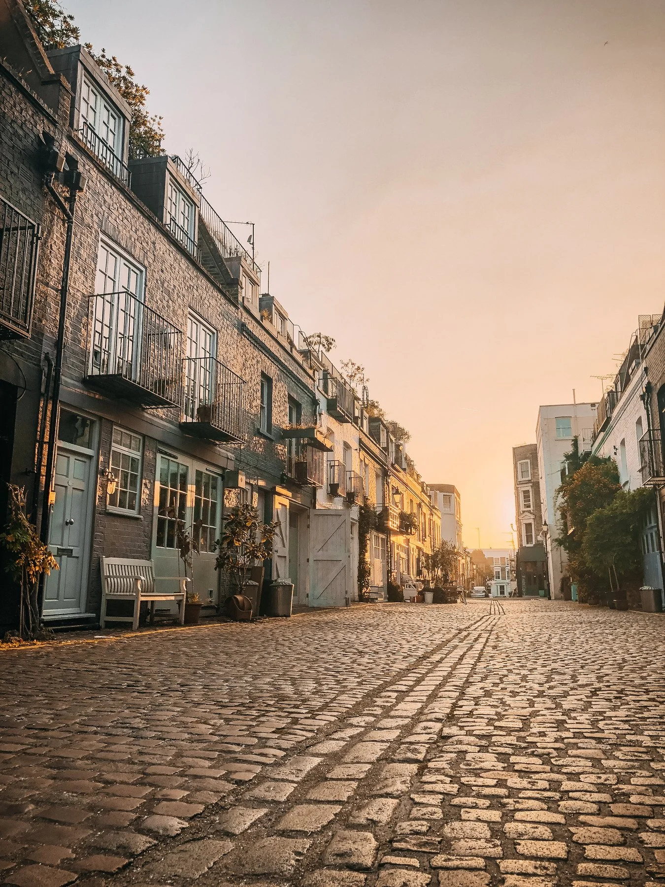 A cobblestone street in a residential neighborhood during sunset with multi-story buildings on either side, some with balconies and plants.