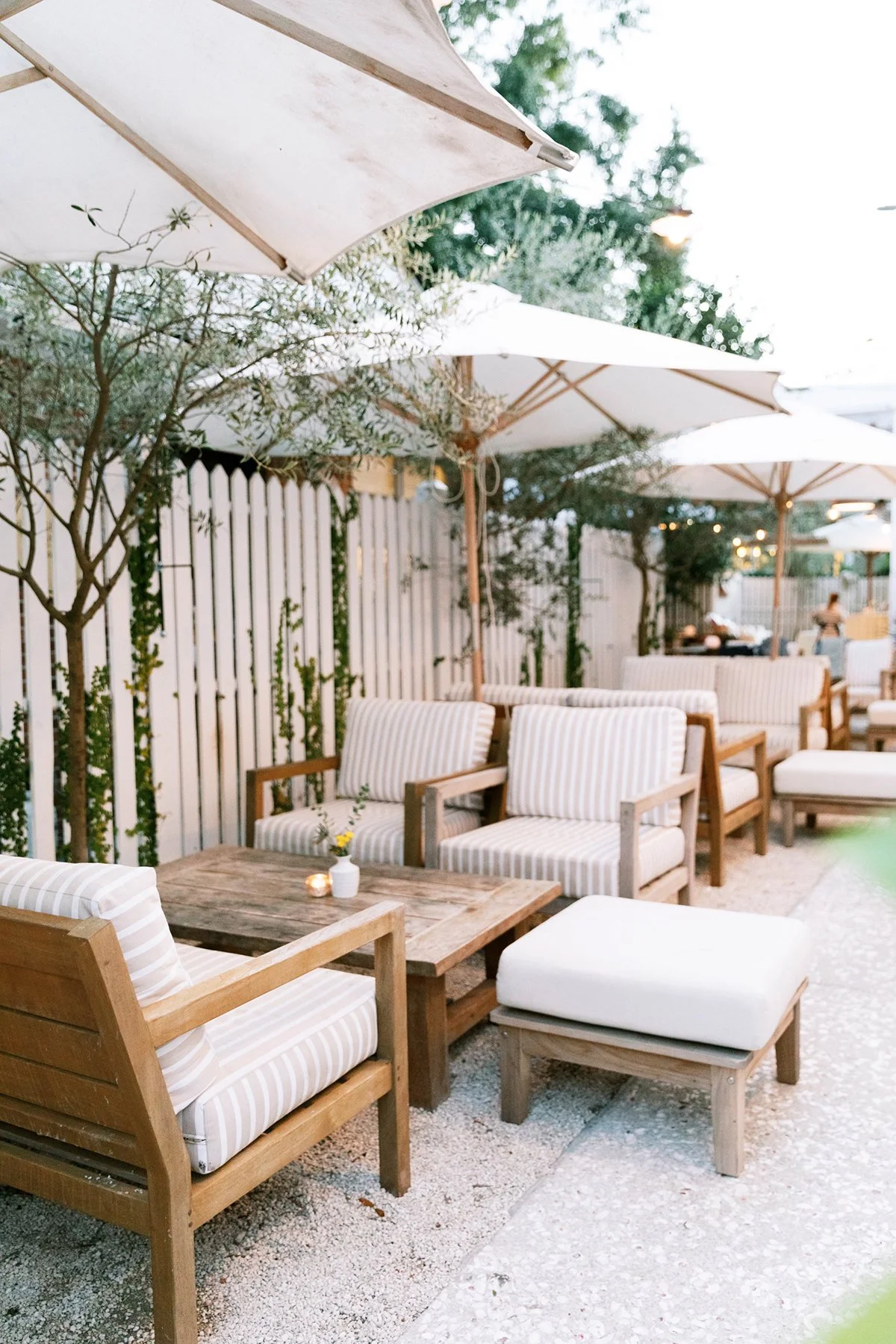 Outdoor patio with wooden furniture, striped cushions, white umbrellas, small flowers in vases, and trees, set for dining or relaxation.