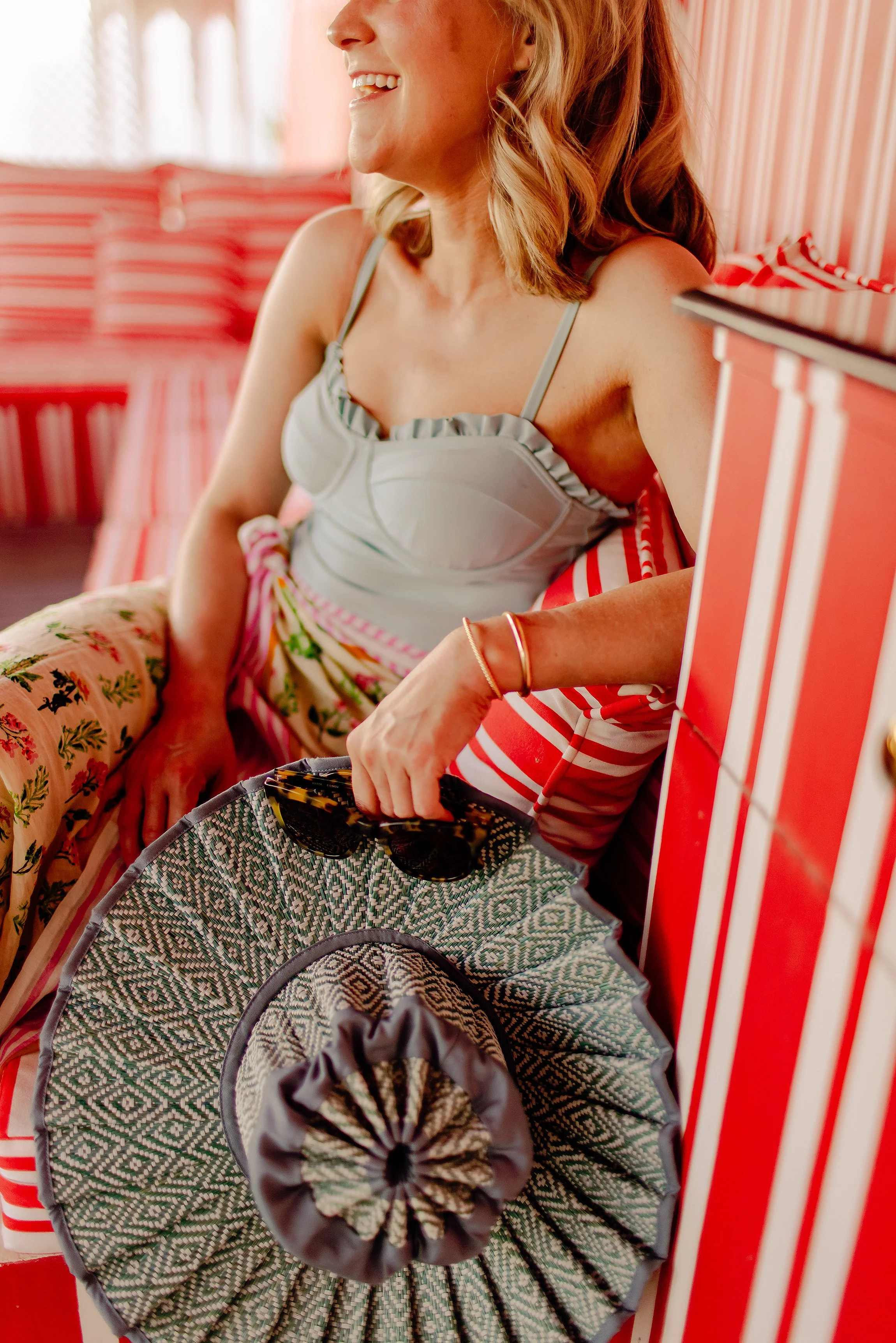 Woman sitting on a red and white striped patio chair, holding a patterned hat and sunglasses, smiling, in a bright, colorful outdoor setting.