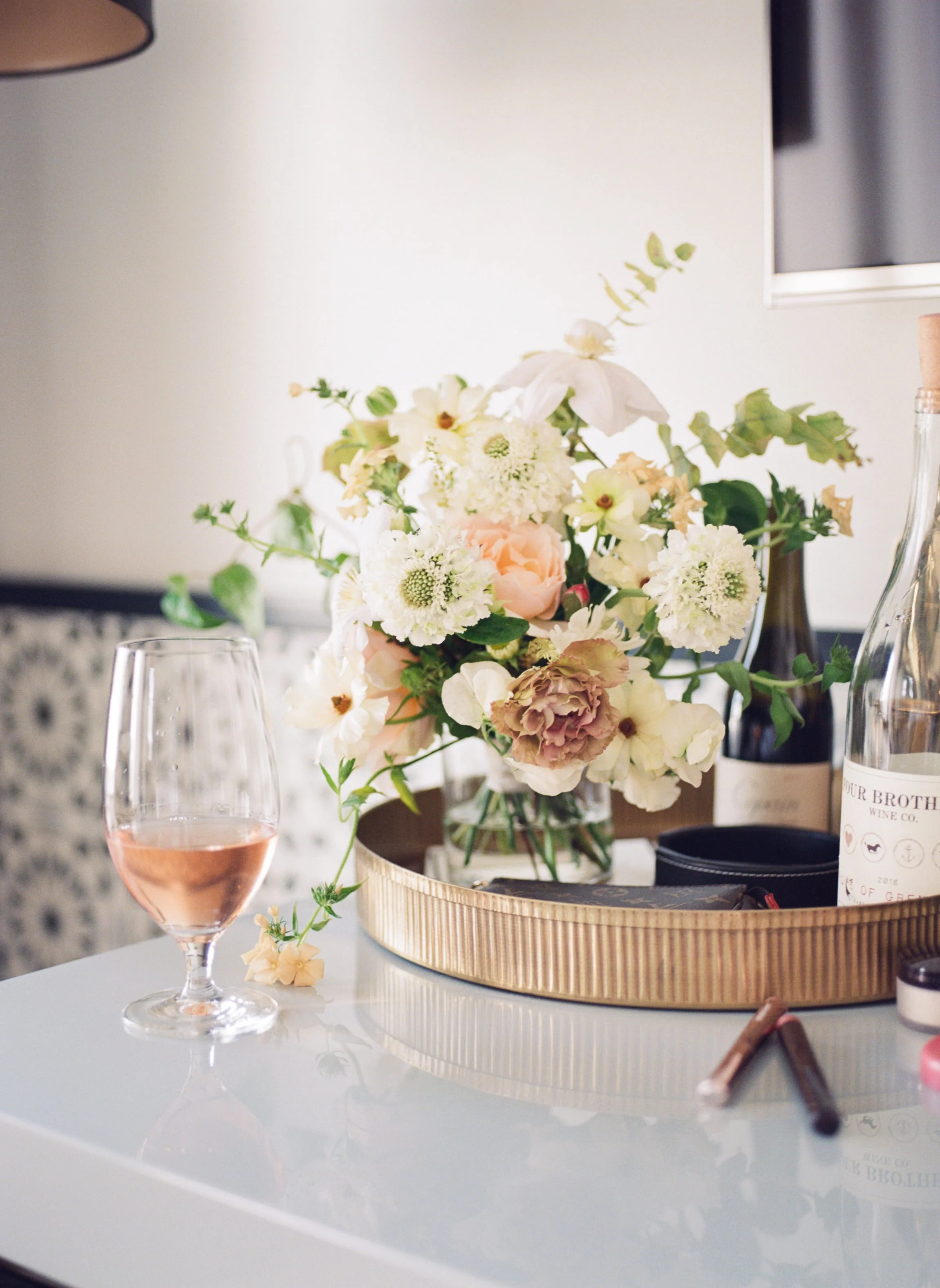 A table with a pink wine glass, a bouquet of white and pastel flowers in a glass vase, a bottle of wine, a water bottle, and makeup items.
