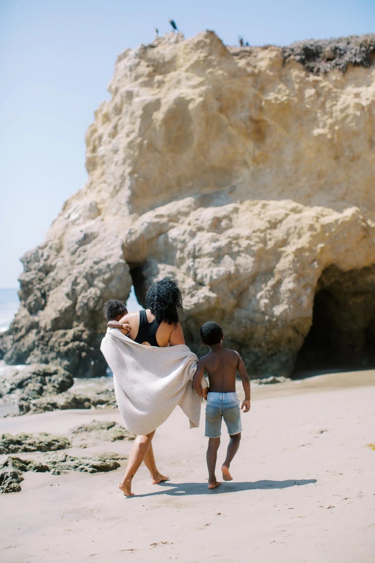 A woman walking on the beach with two children, one in her arms and the other holding her hand, near large rocks and cave openings.