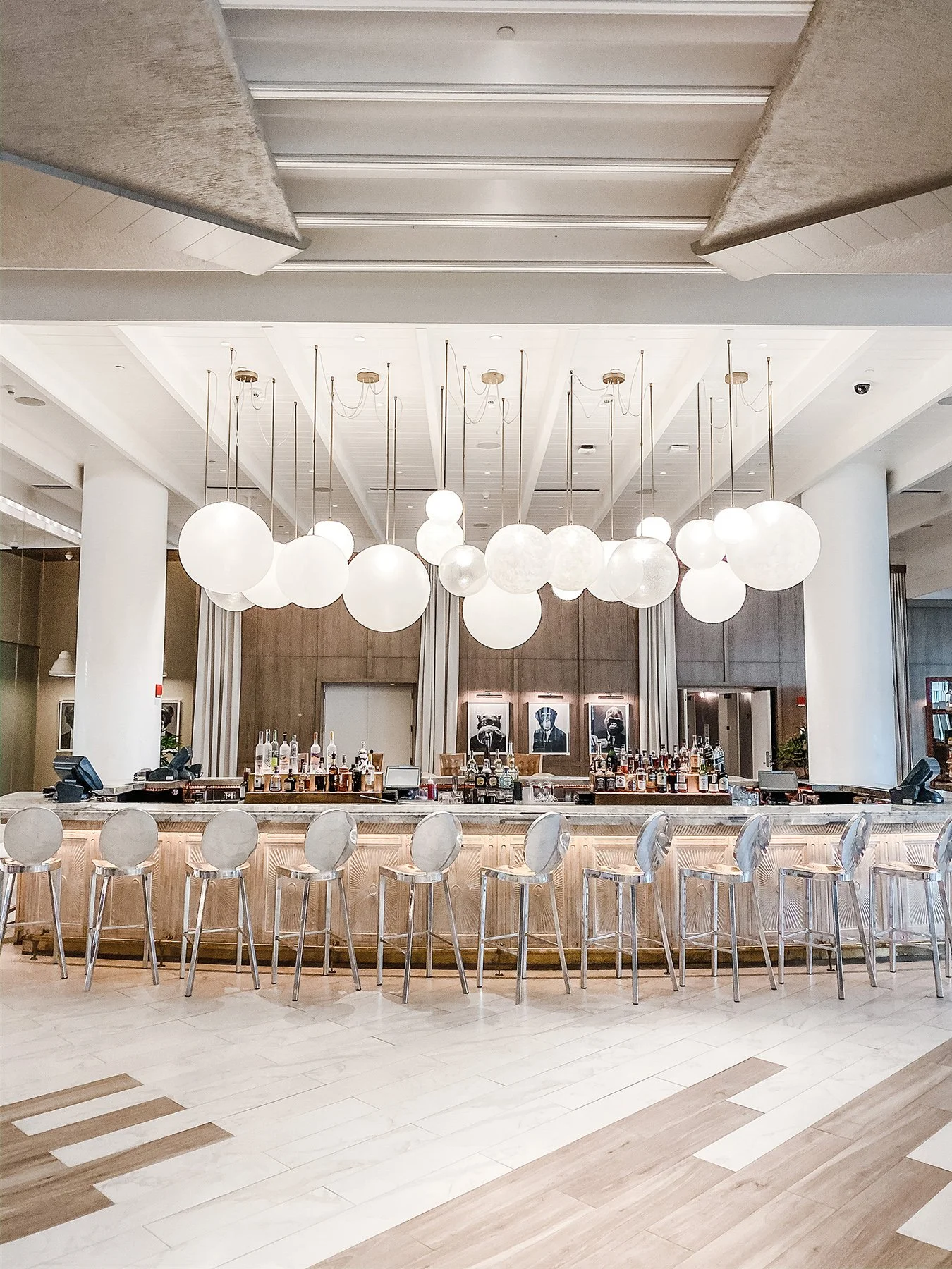 A modern hotel bar with white spherical hanging lights, a curved marble countertop, and light wood paneling behind the bar with framed pictures.