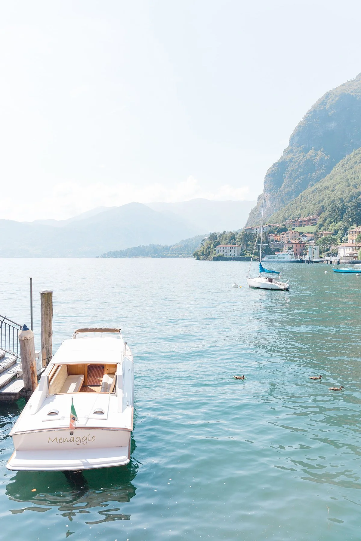 A peaceful lakeside scene with a white boat named 'Menaggio' docked at a pier, surrounded by water with ducks swimming nearby. In the background, there are sailboats on the lake, a hillside with houses, and green mountains under a partly cloudy sky.