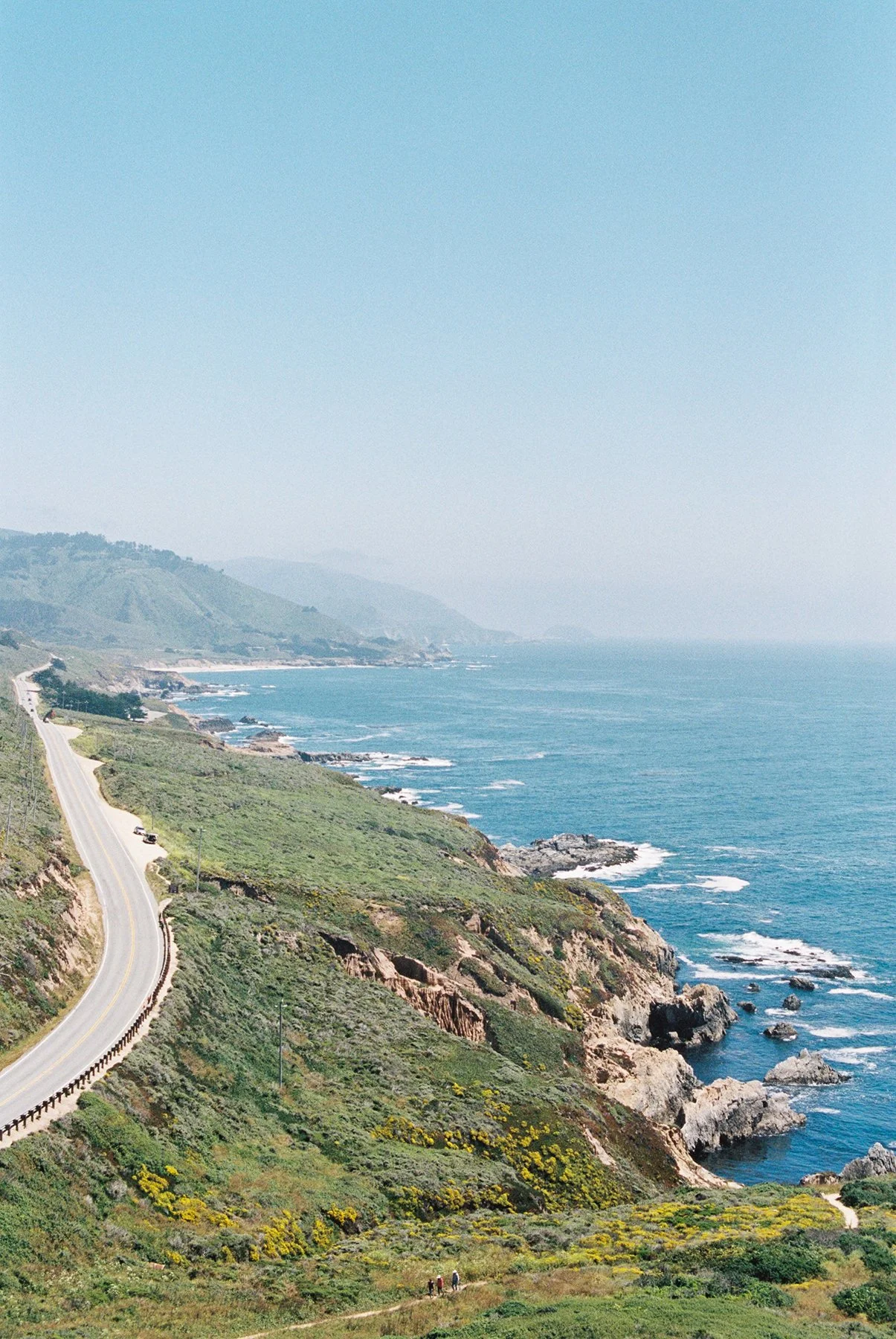 Scenic view of a coastal highway winding along green cliffs overlooking the ocean with rocks, waves, and distant mountains under a clear blue sky.