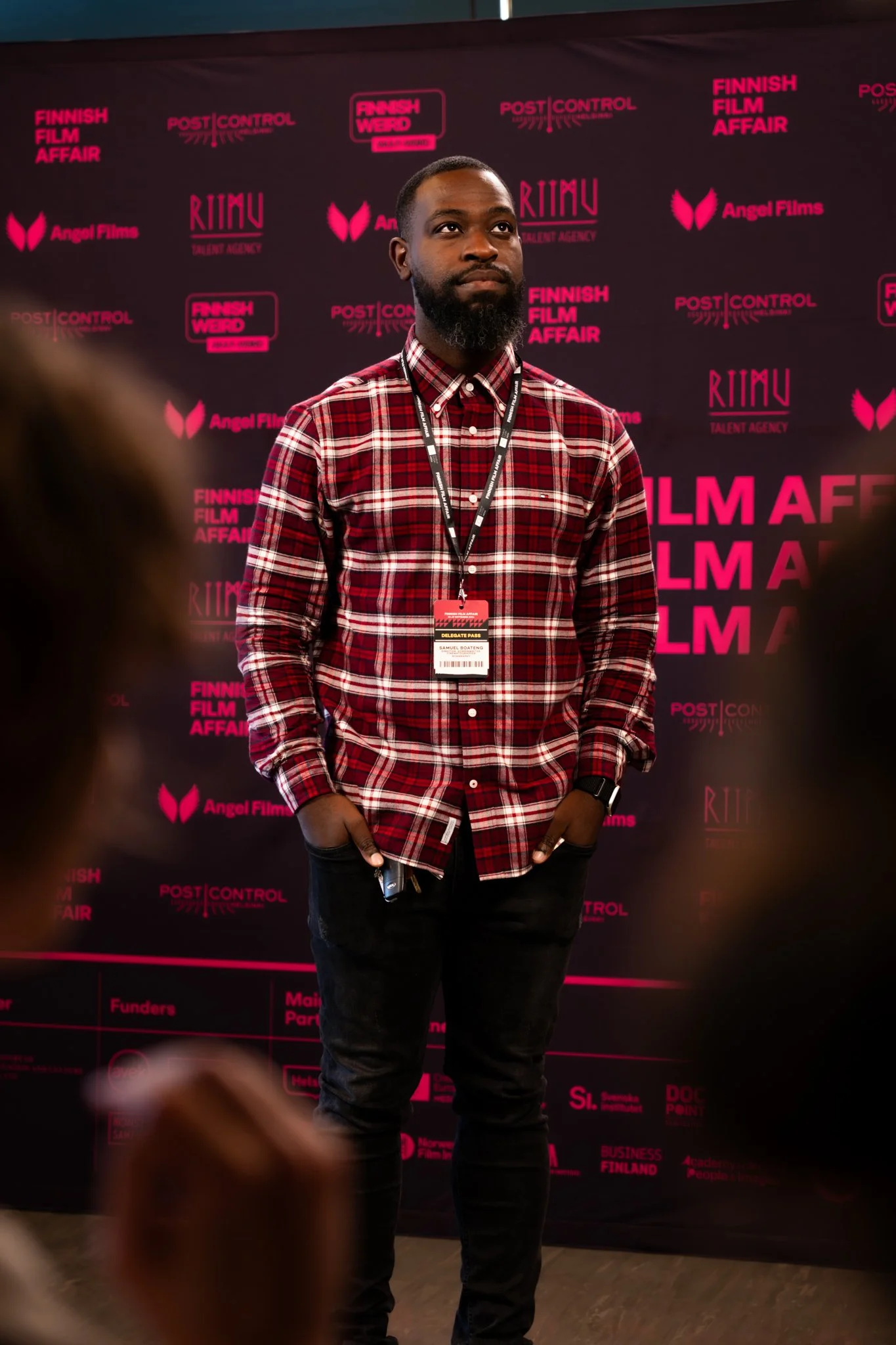 A man in a red plaid shirt standing in front of a black backdrop with pink text and logos at a film event.