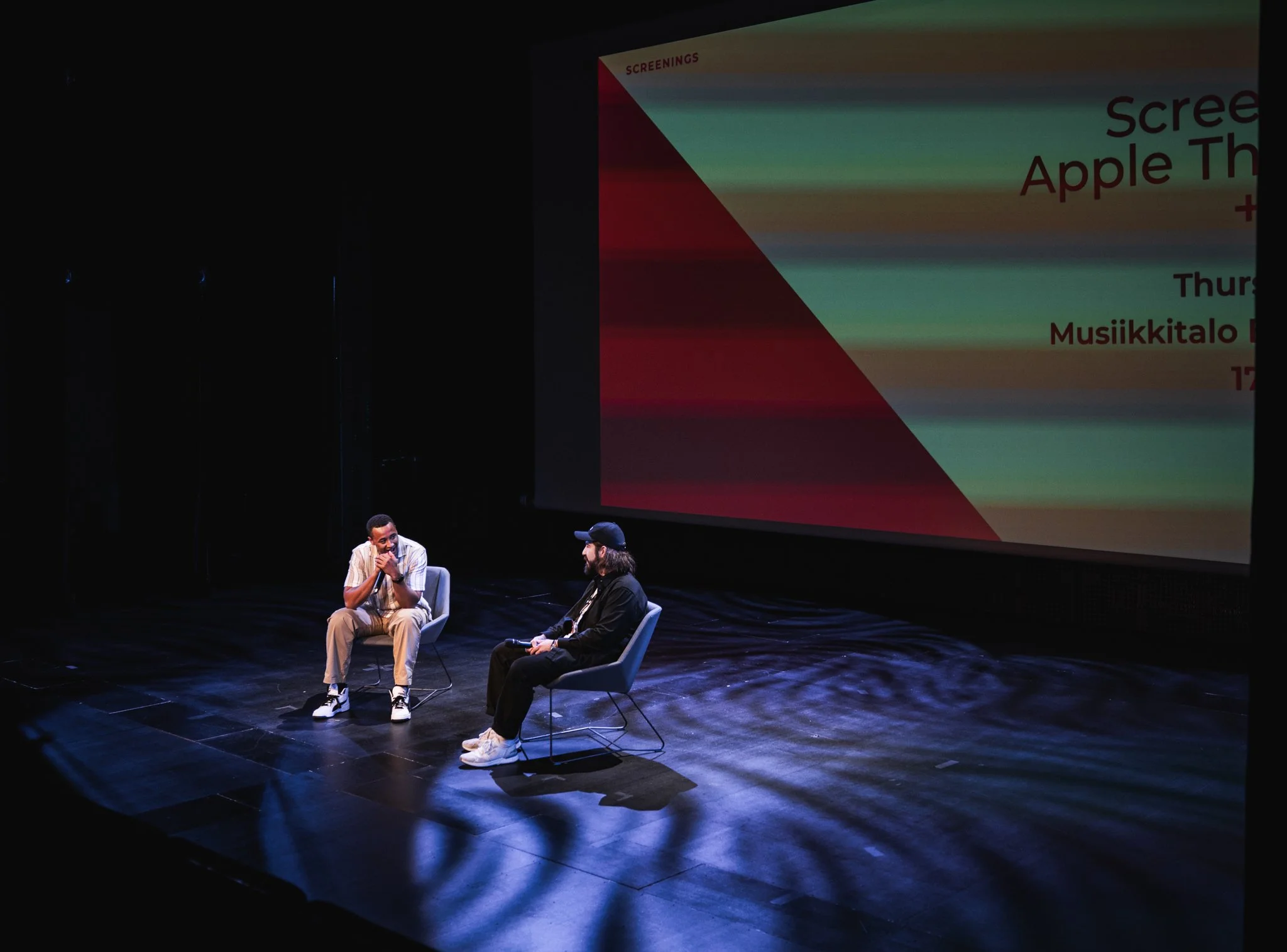 Two men sitting on stage chairs having a conversation with a large screen behind them displaying an event announcement.