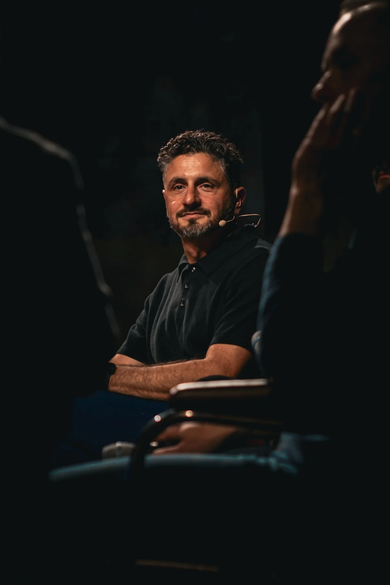 A man with curly hair and a beard wearing a black polo shirt, sitting at a panel or discussion with others, looking towards the camera with a slight smile.