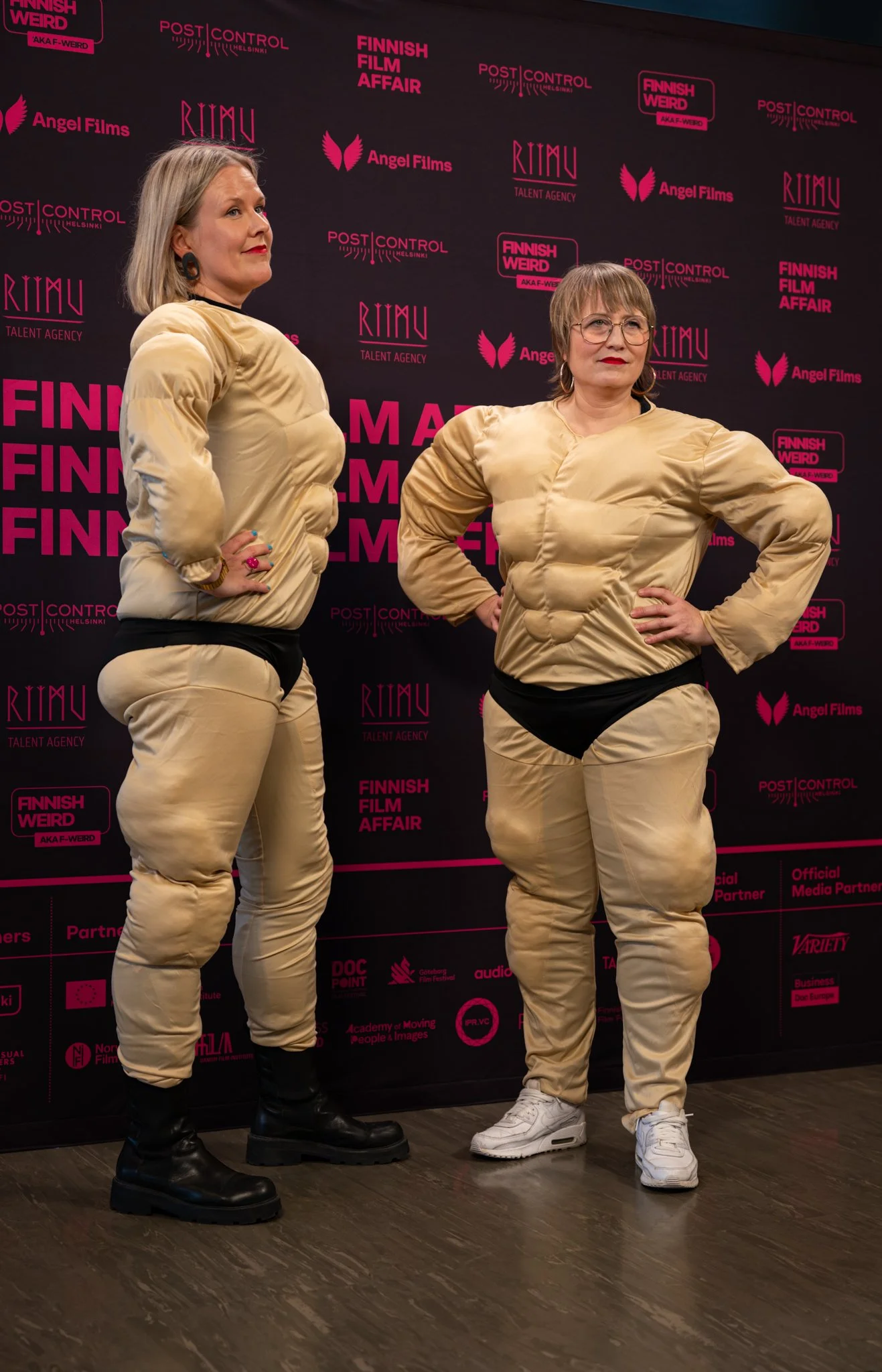 Two women dressed in padded beige outfits with black and white bottoms, posing with hands on hips in front of a black and pink backdrop at the Finnish Film Affair event.